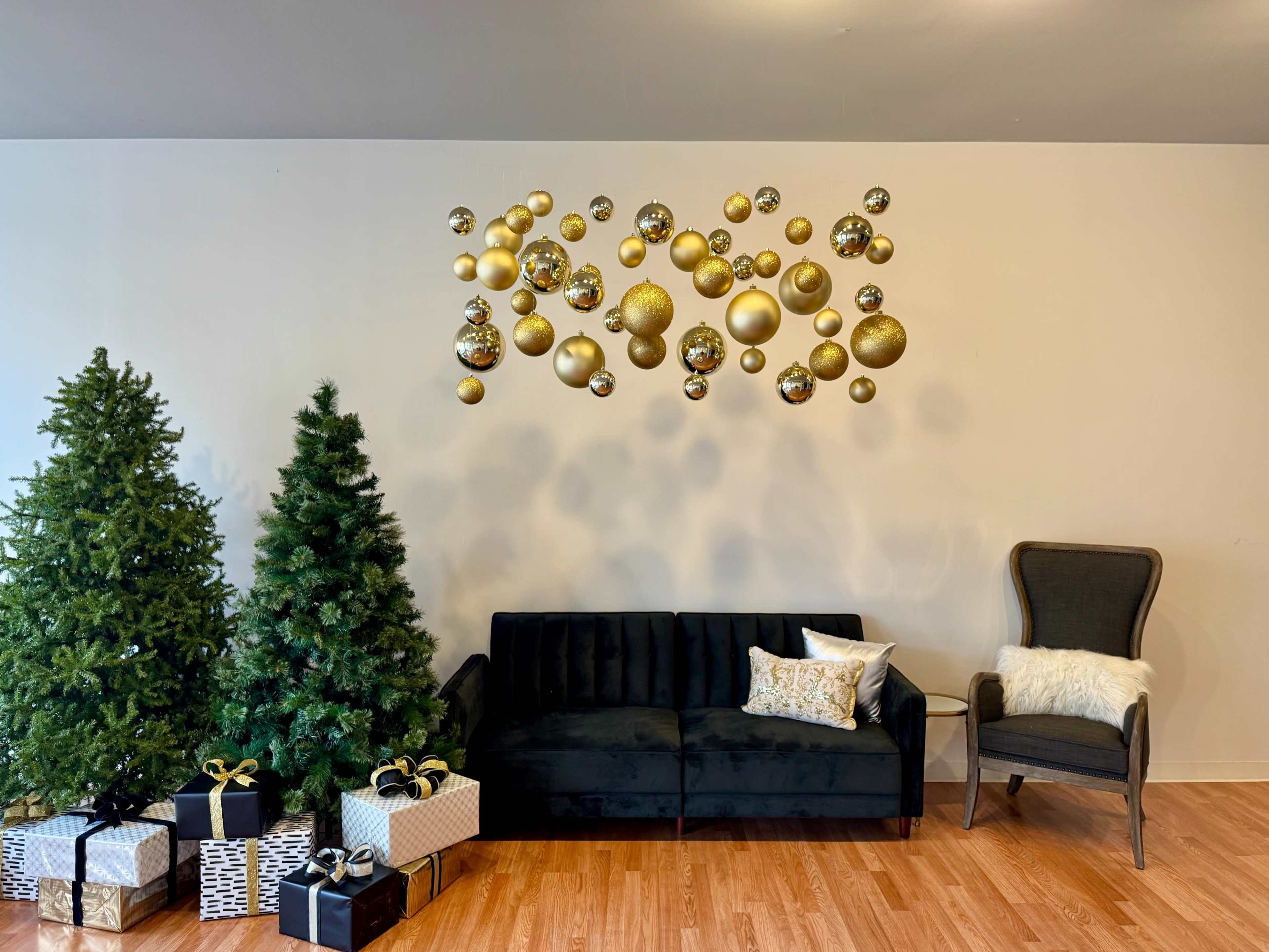 The image features a living room with two Christmas trees, a dark sofa, and a wall adorned with gold ornaments, complemented by gifts and a chair.
