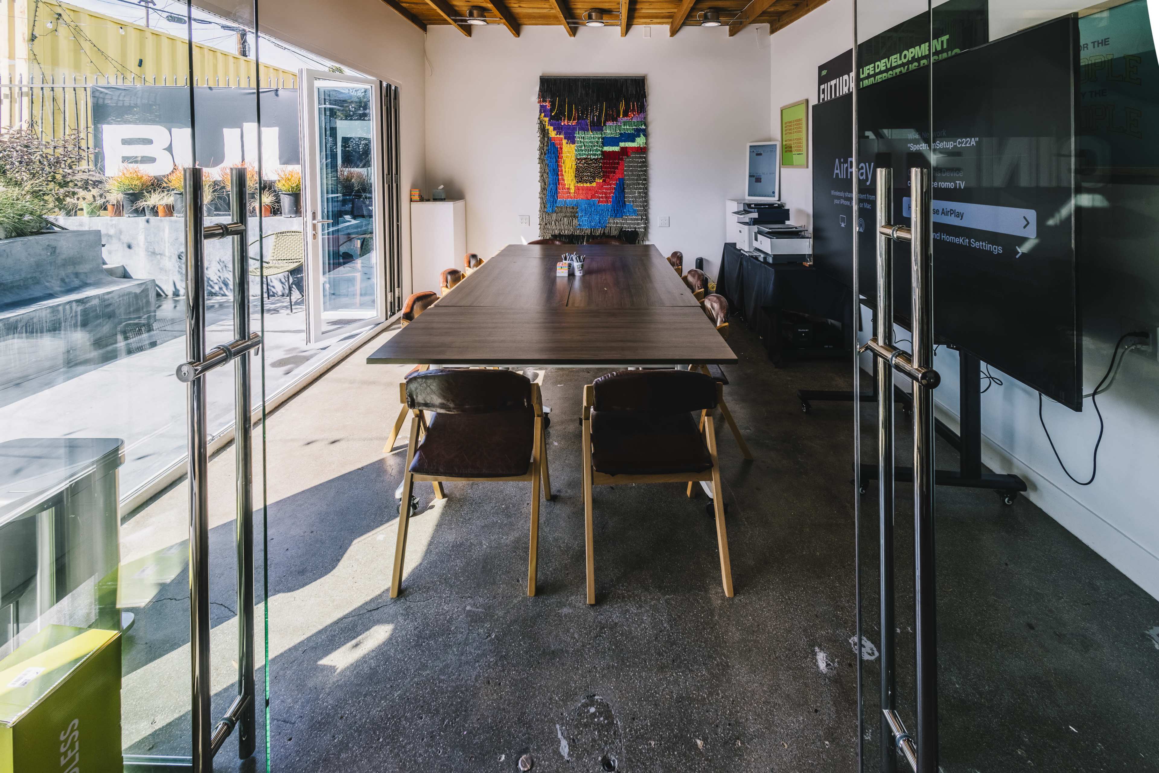 The image shows a modern conference room with a large wooden table surrounded by chairs, glass doors leading outside, and a wall-mounted screen displaying information.