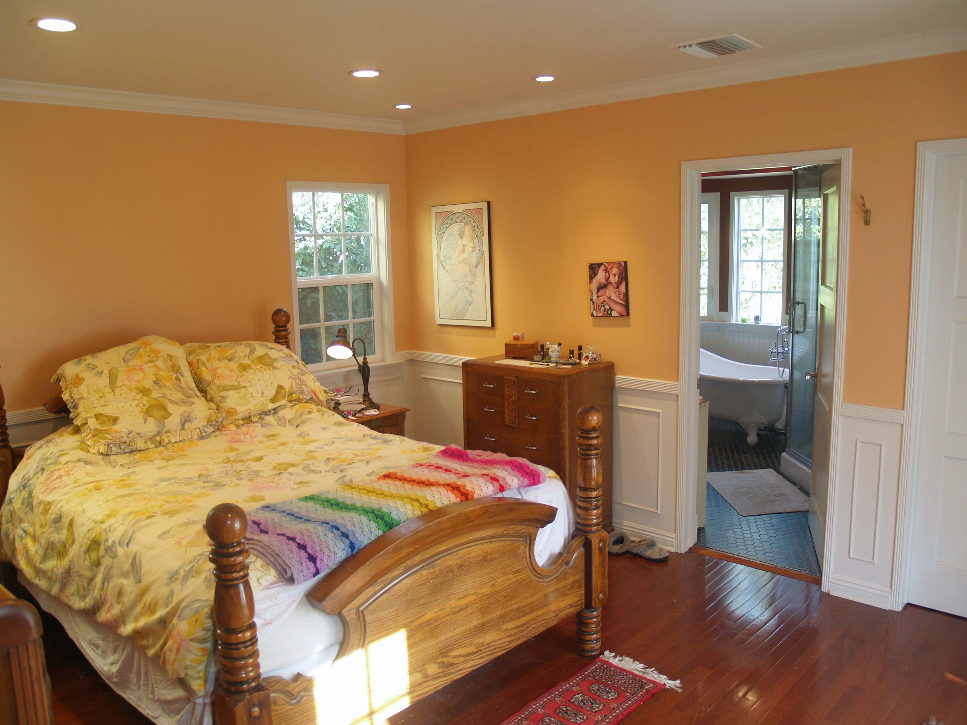 The bedroom features a wooden bed with a colorful blanket, hardwood flooring, and an adjoining bathroom visible through an open door.