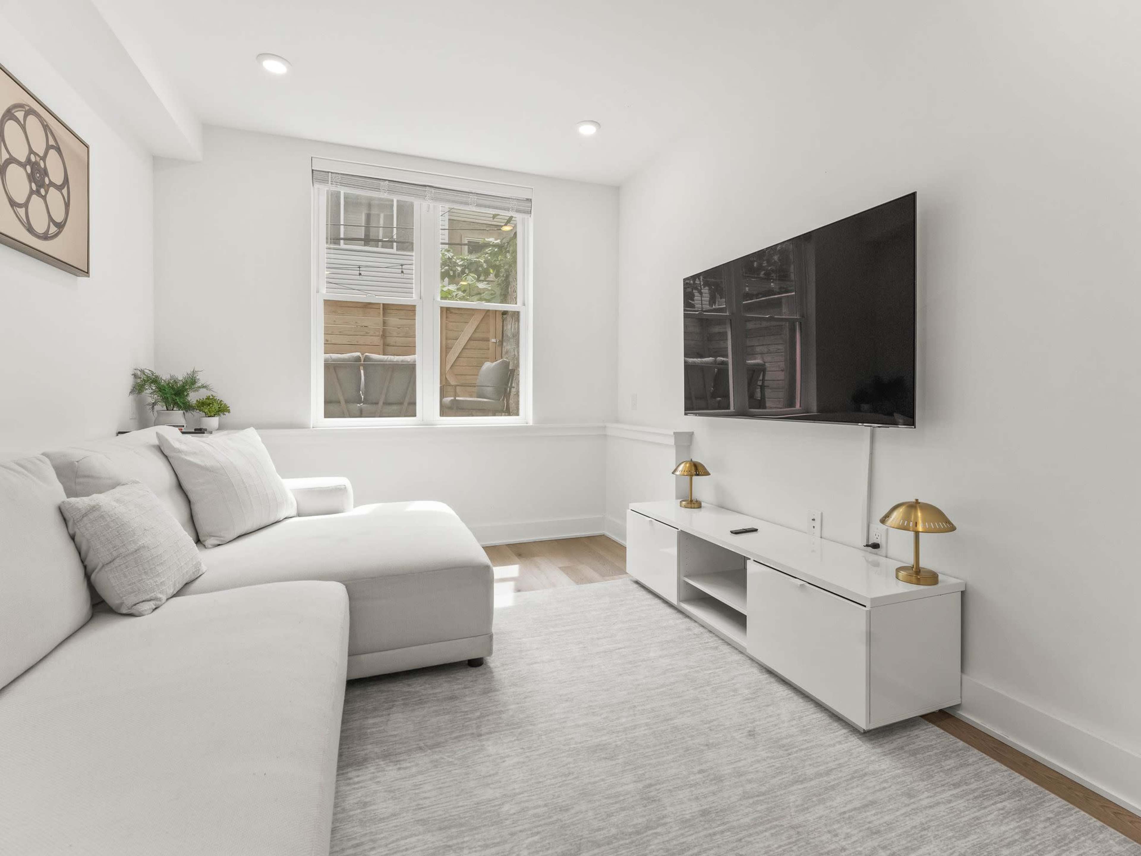 The image shows a minimalist living room featuring a white sectional sofa, a flat-screen TV mounted on the wall, and a sleek white media console with two gold lamps.