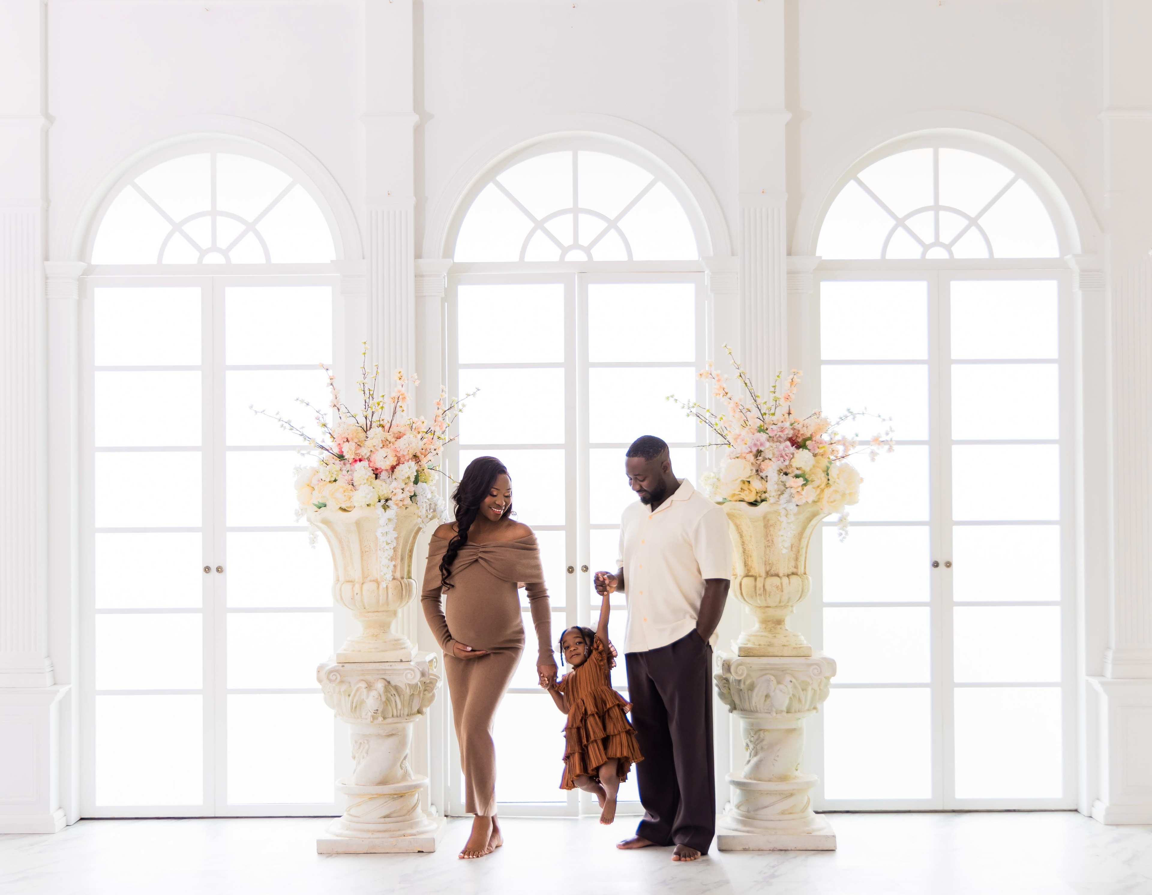 A pregnant woman and a man walk hand-in-hand with a girl in a brown dress, framed by large floral arrangements near tall windows.