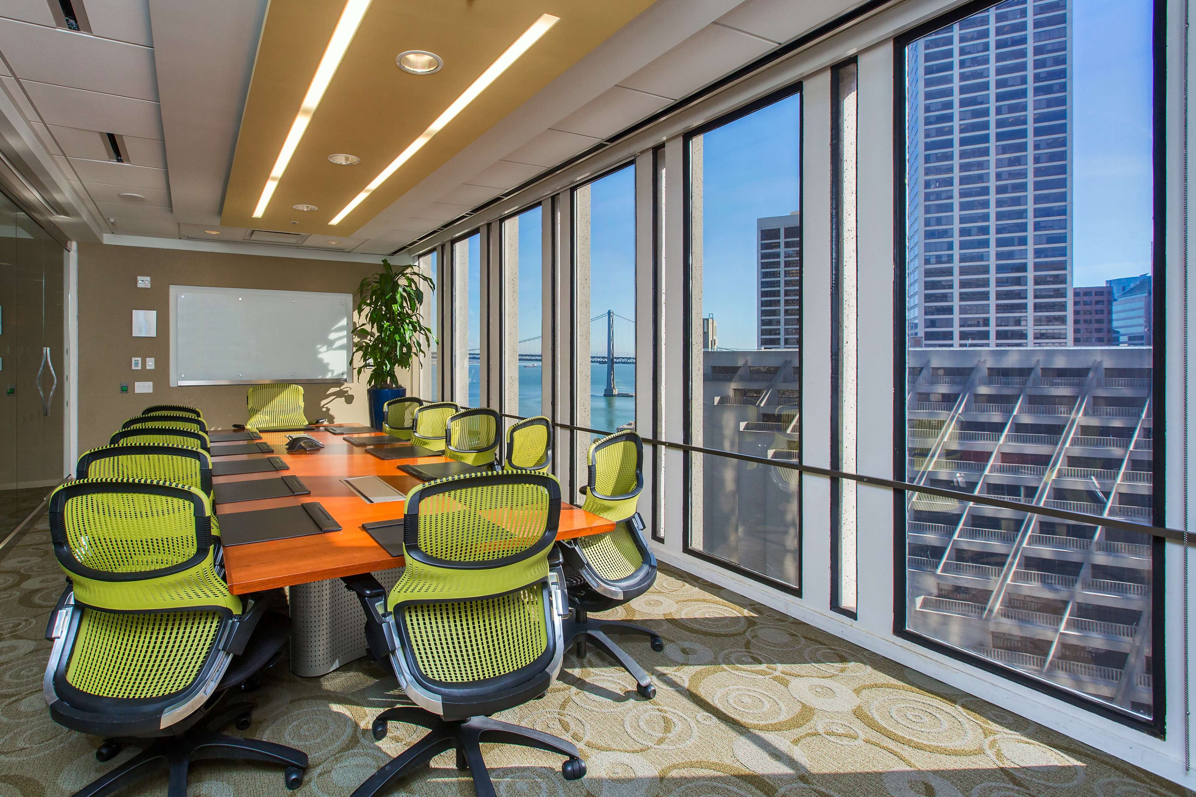A modern conference room features a large wooden table surrounded by green office chairs and floor-to-ceiling windows overlooking a cityscape and water.