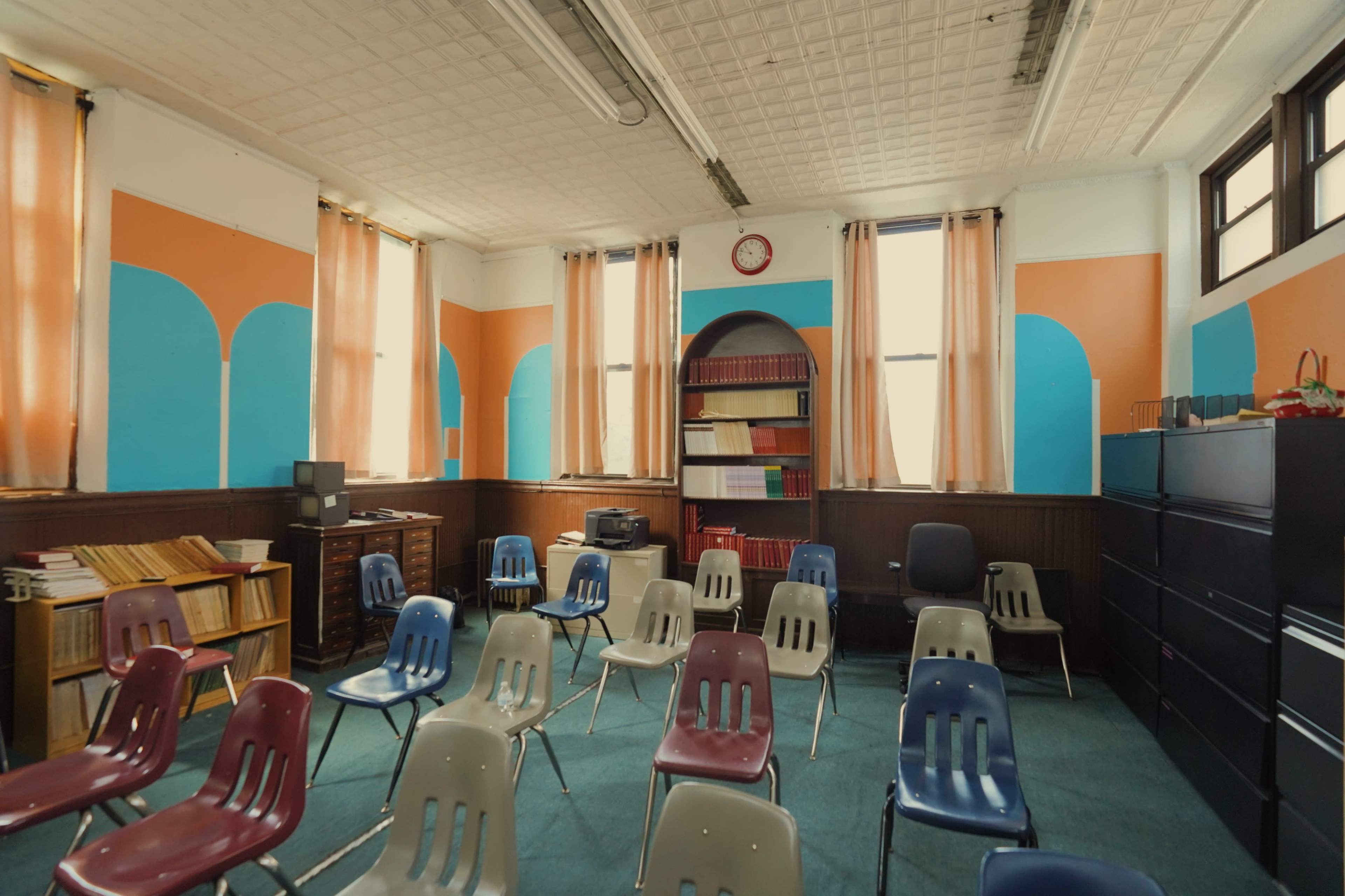 The image depicts a classroom with rows of plastic chairs, a wooden bookshelf, and brightly colored walls.