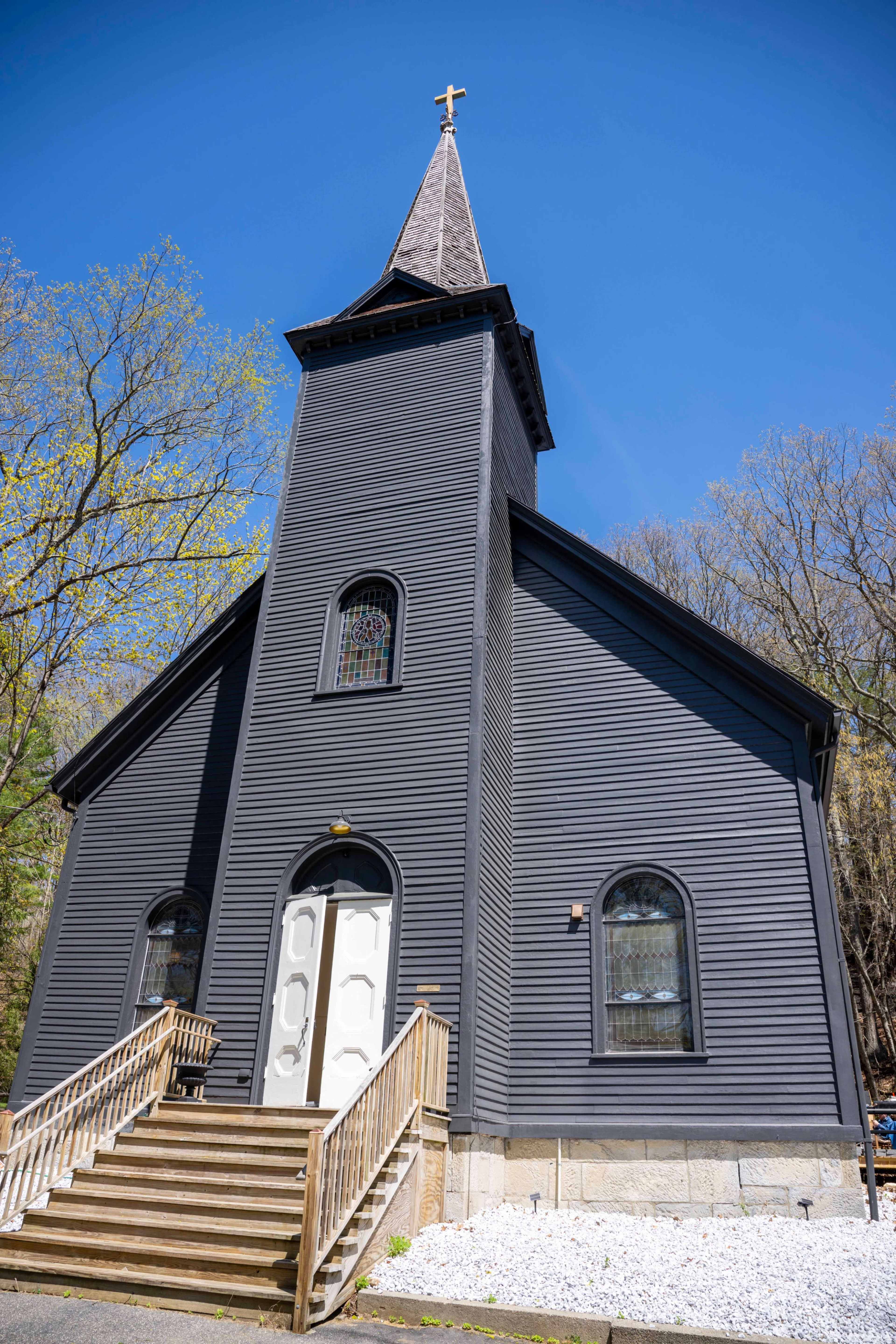 The image features a gray wooden church with a pointed steeple, a white entrance door, and a set of wooden steps leading to the front.