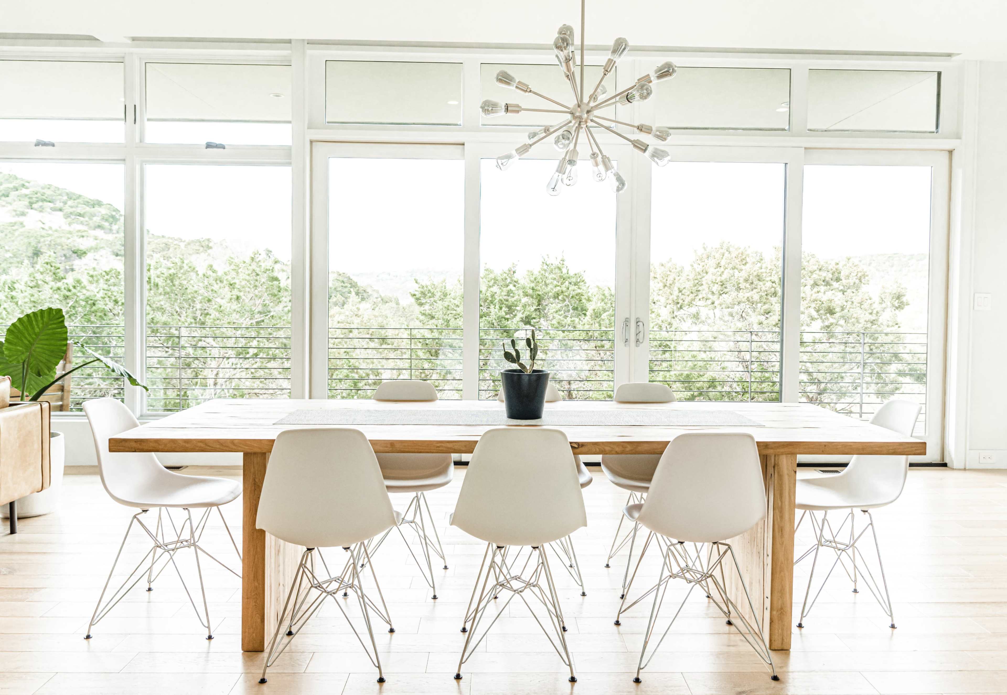A long wooden dining table surrounded by white chairs is set against large windows overlooking greenery.