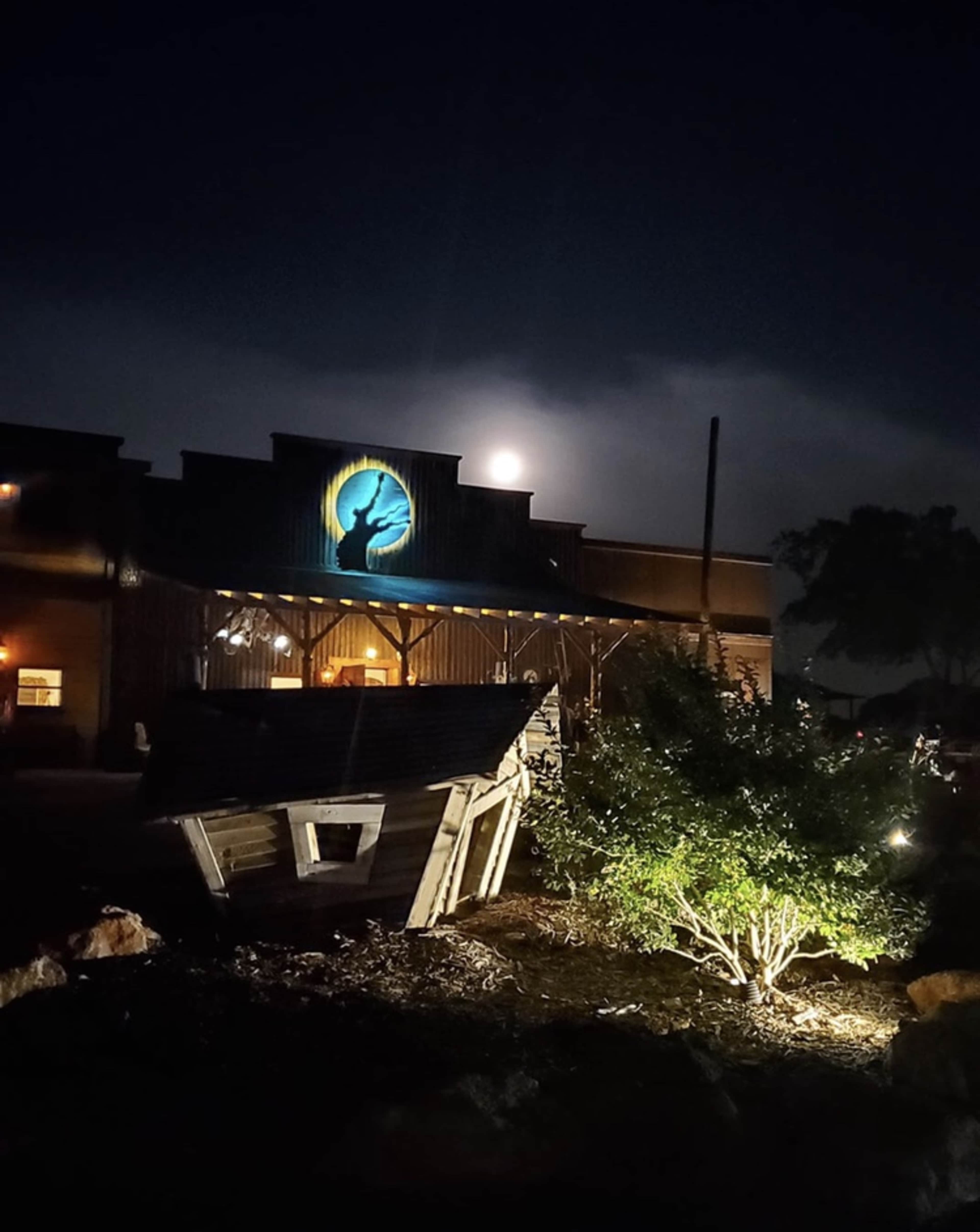 A full moon illuminates the facade of a rustic building, while a wooden sign and shrubbery are lit by ground lights in the foreground.