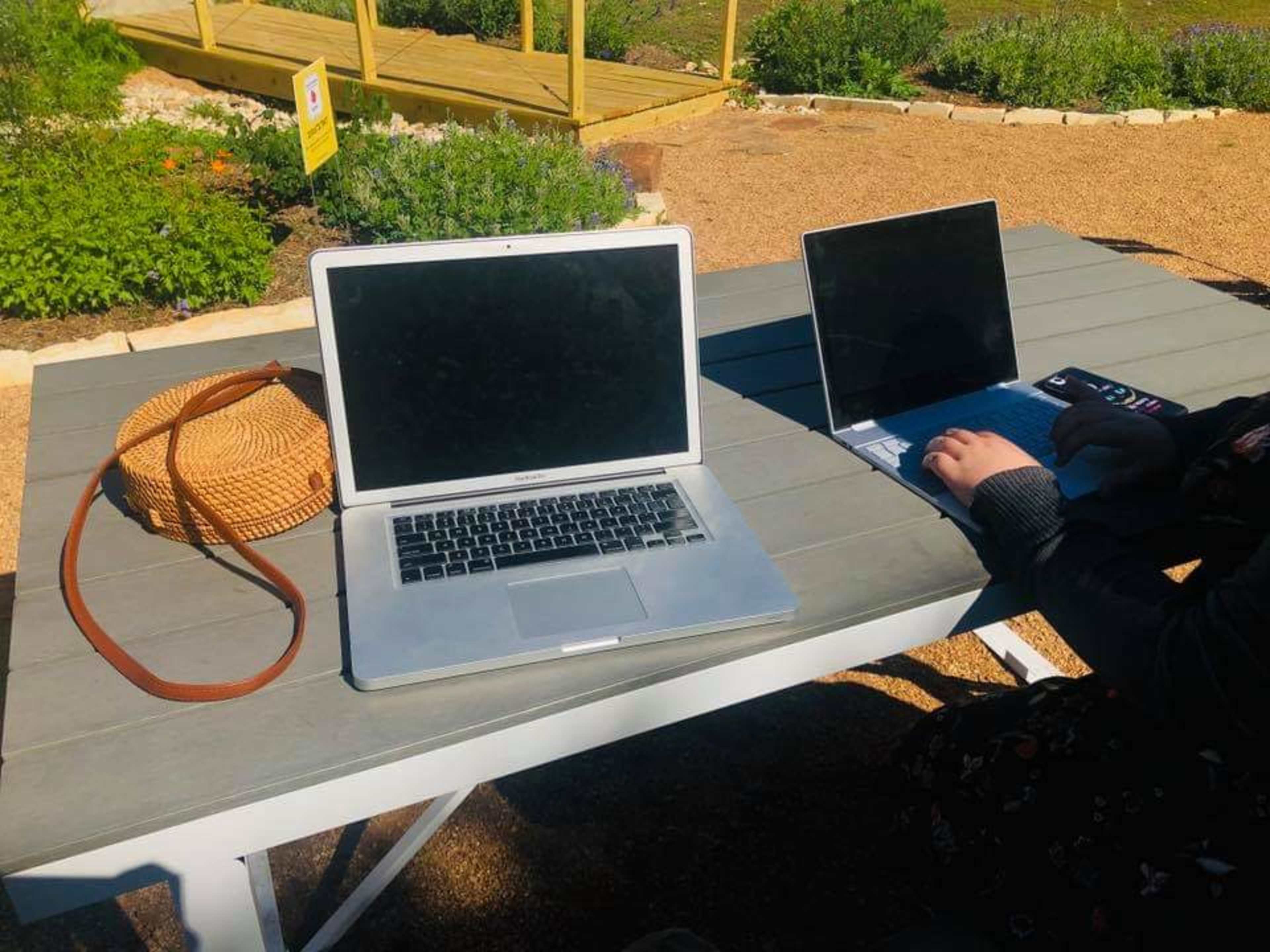 A wooden table in a garden setting holds two laptops and a woven bag.