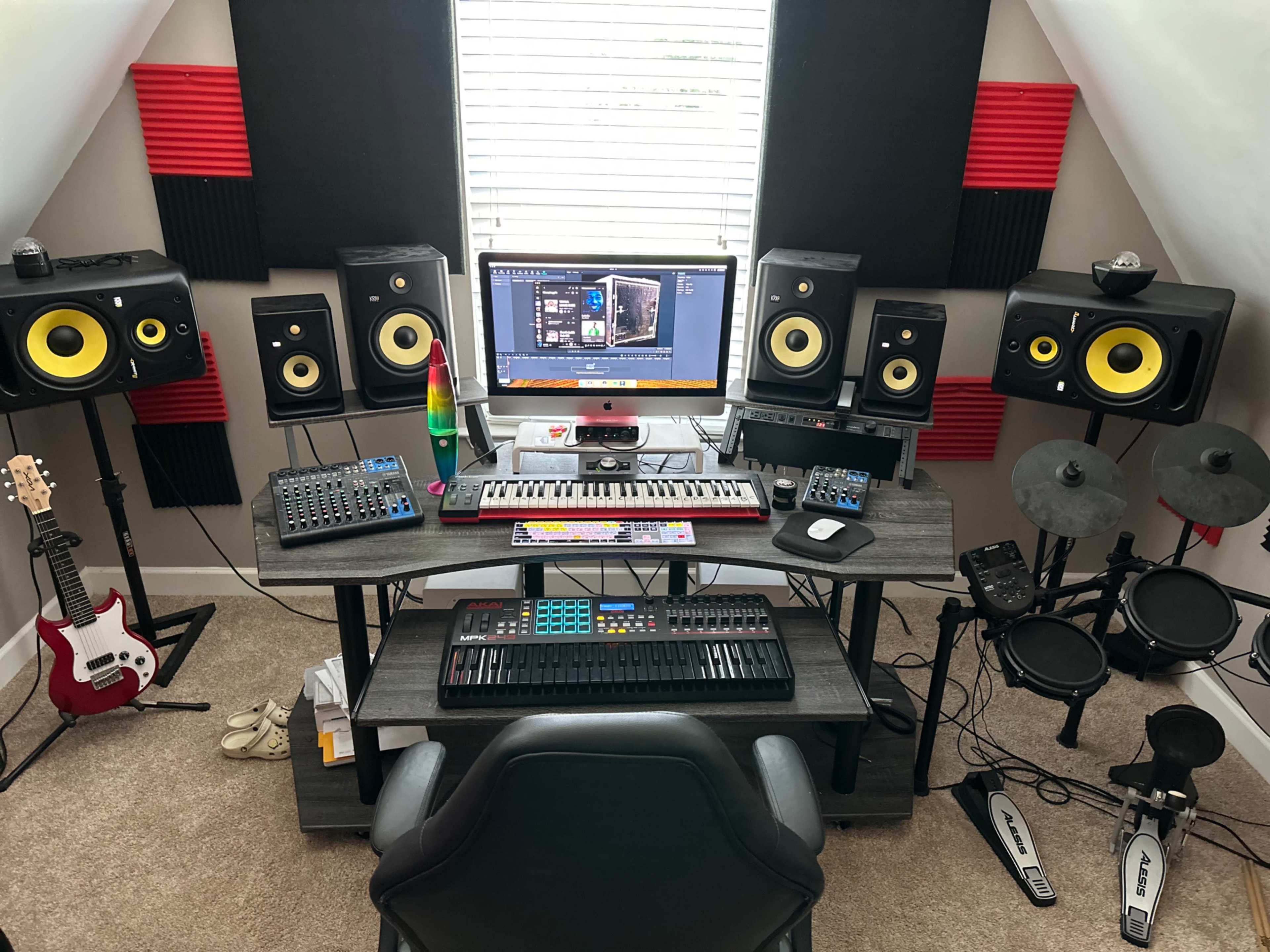 The image shows a music production setup with a central computer monitor, studio monitors, a MIDI keyboard, electronic drum pads, and various musical instruments arranged on a desk.