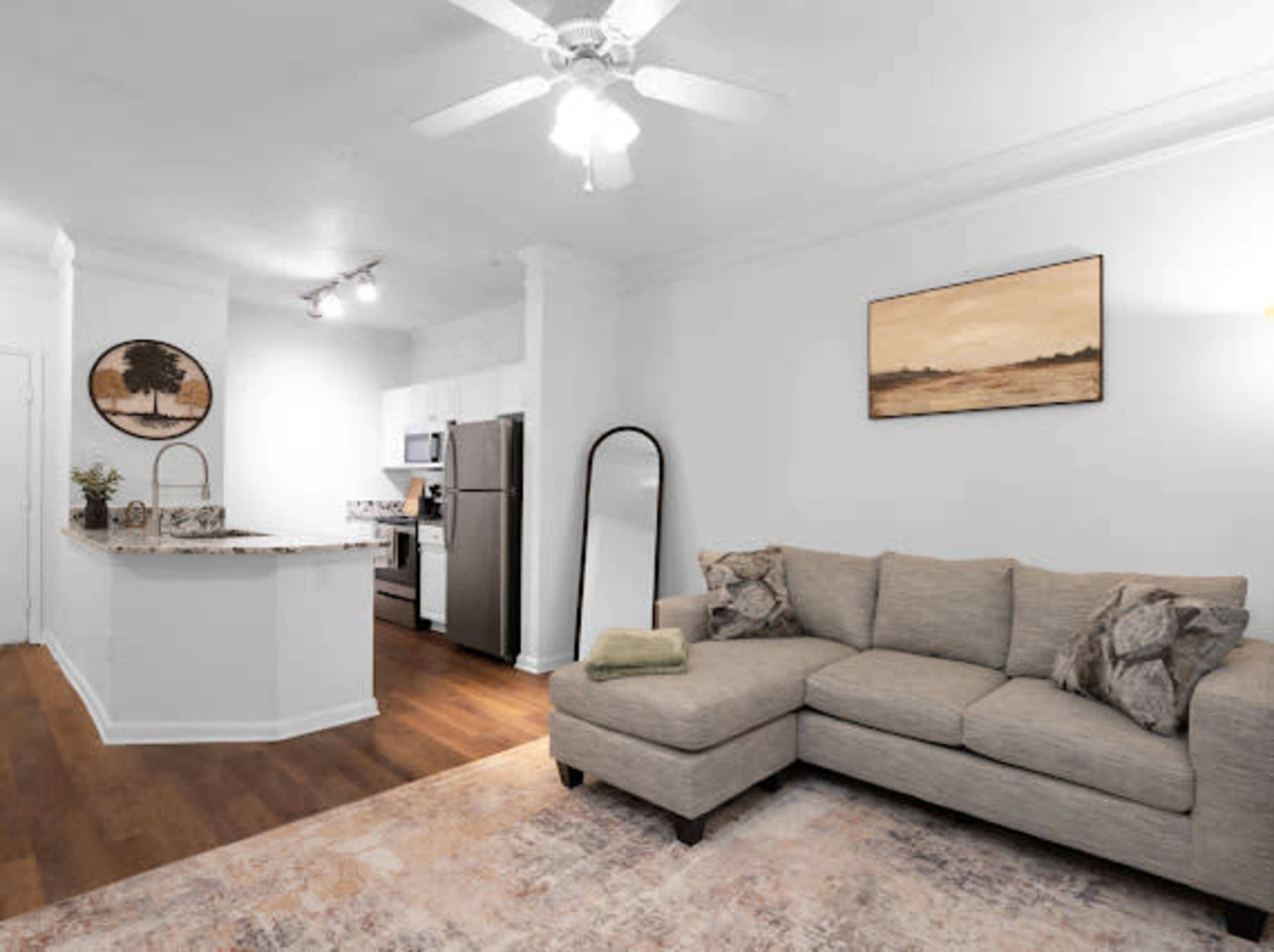 The image shows a contemporary living space featuring a sectional sofa, an open kitchen with modern appliances, and a decorative mirror against a light-colored wall.