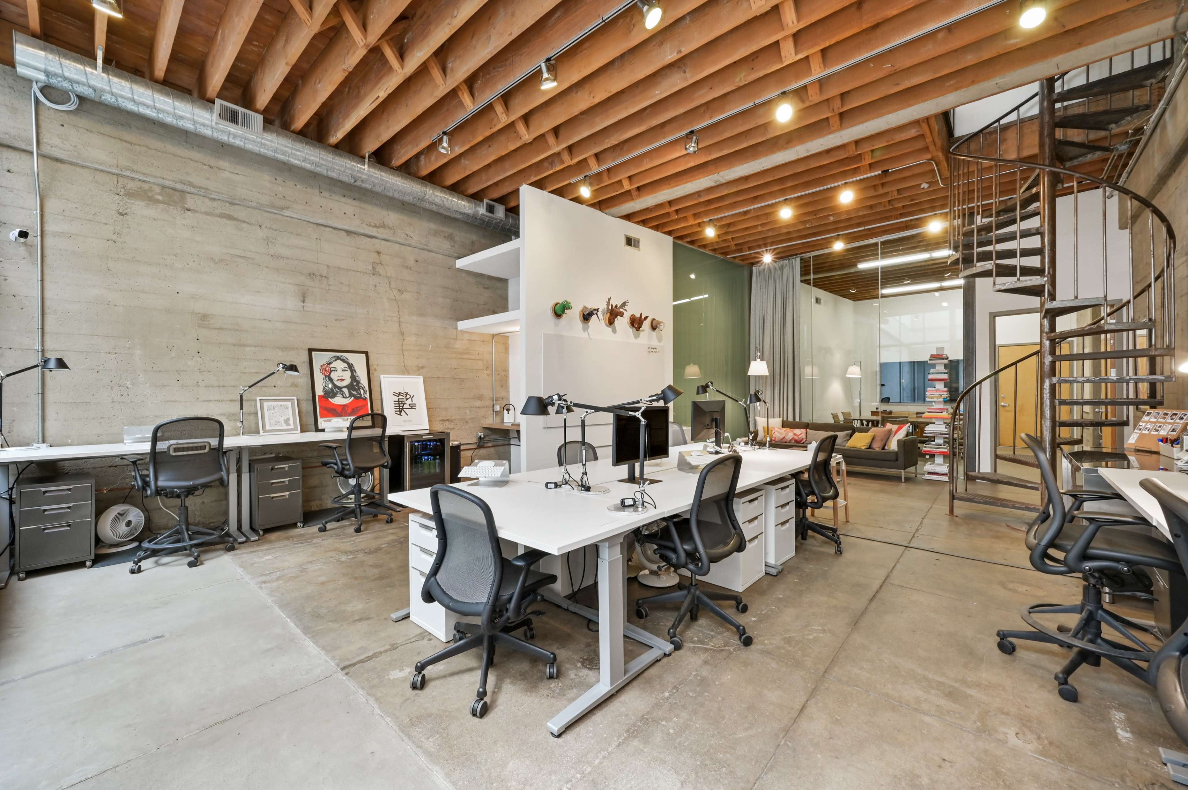 A modern office space with exposed wooden beams, multiple workstations equipped with desks and office chairs, and a spiral staircase leading to an upper level.