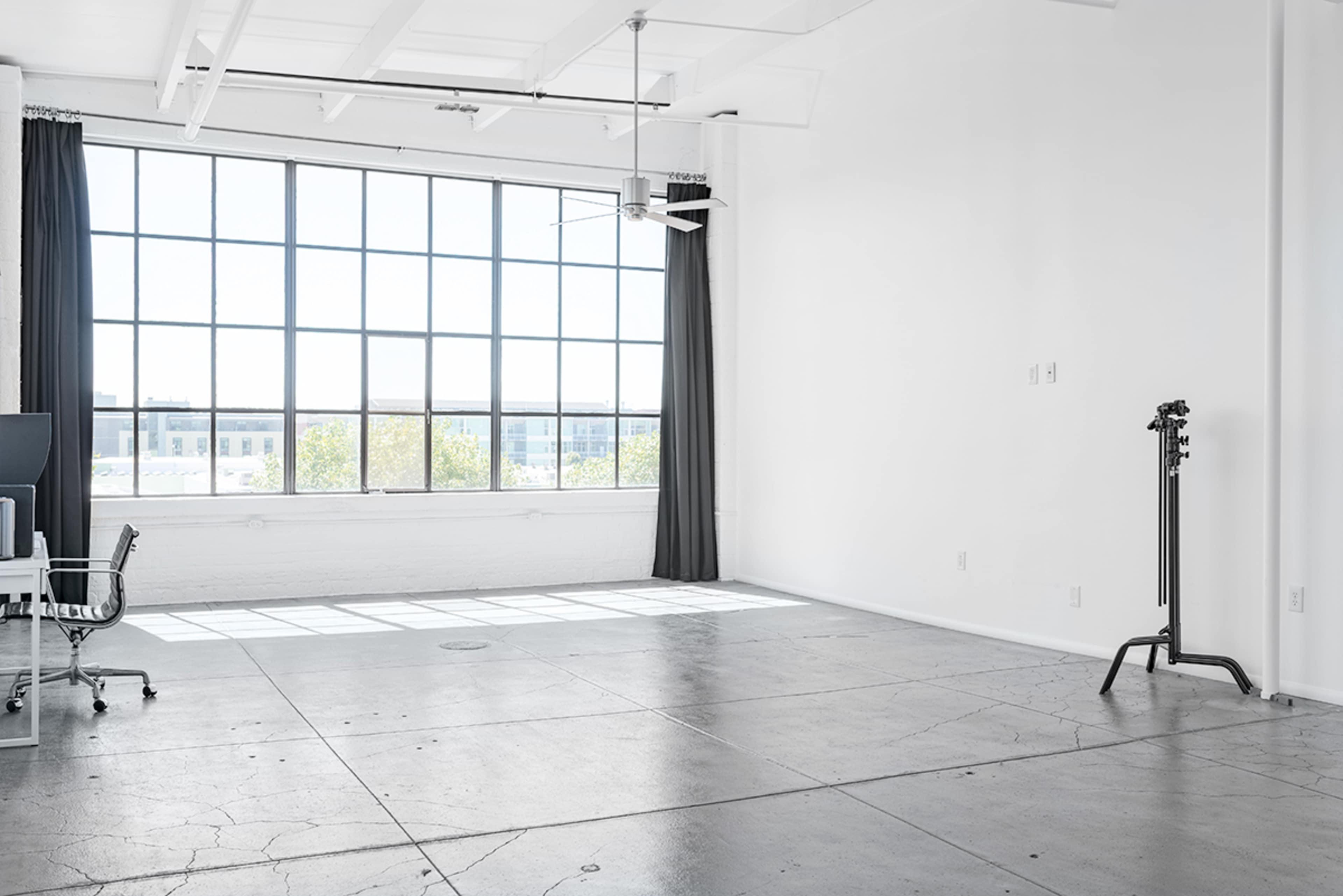A bright, spacious room features a large window with a view, a ceiling fan, and minimal furniture including a chair and a stand.