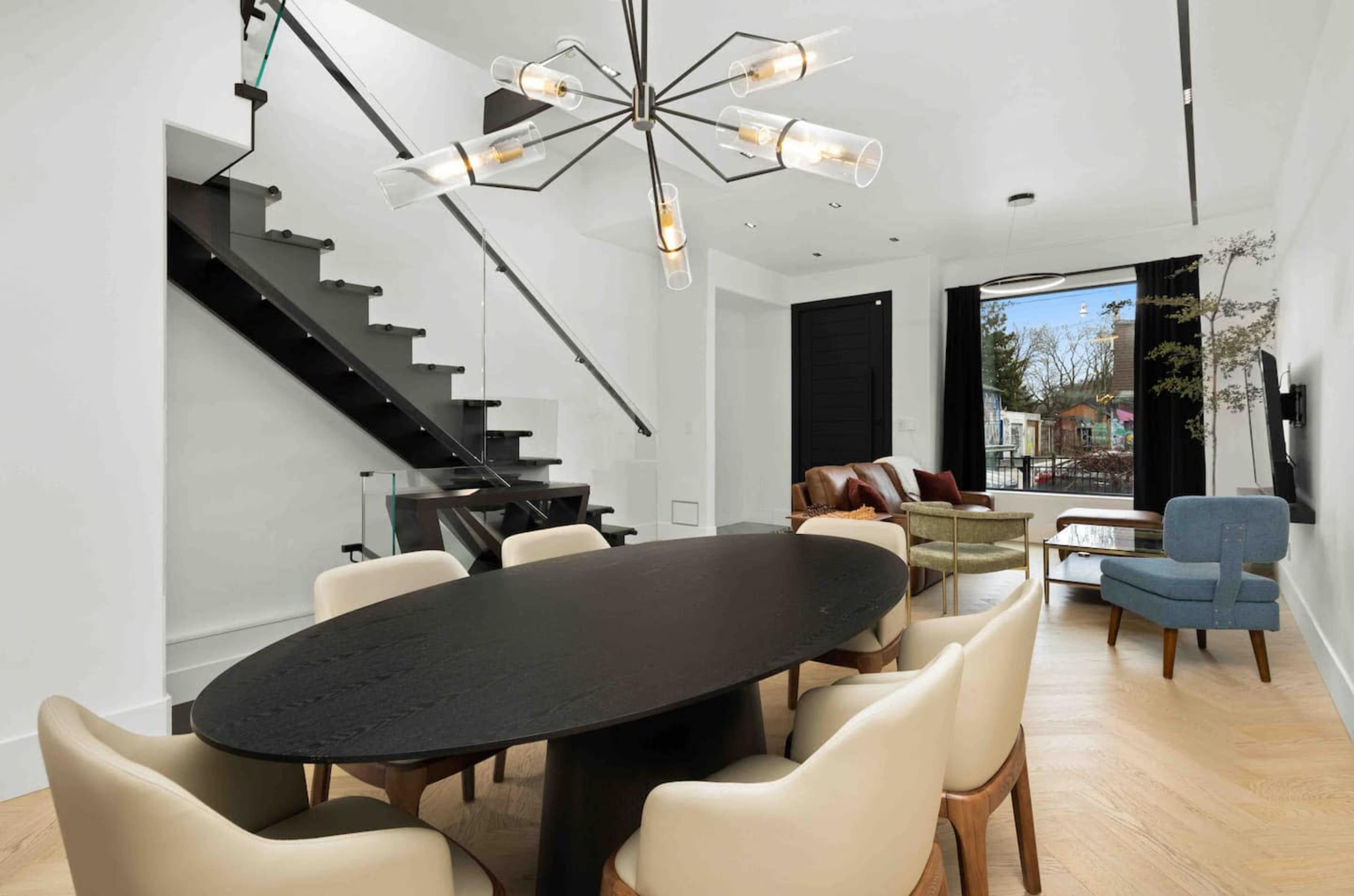 The image shows a modern interior featuring a round black dining table surrounded by beige chairs, a contemporary chandelier overhead, and a staircase in the background leading to an upper level.