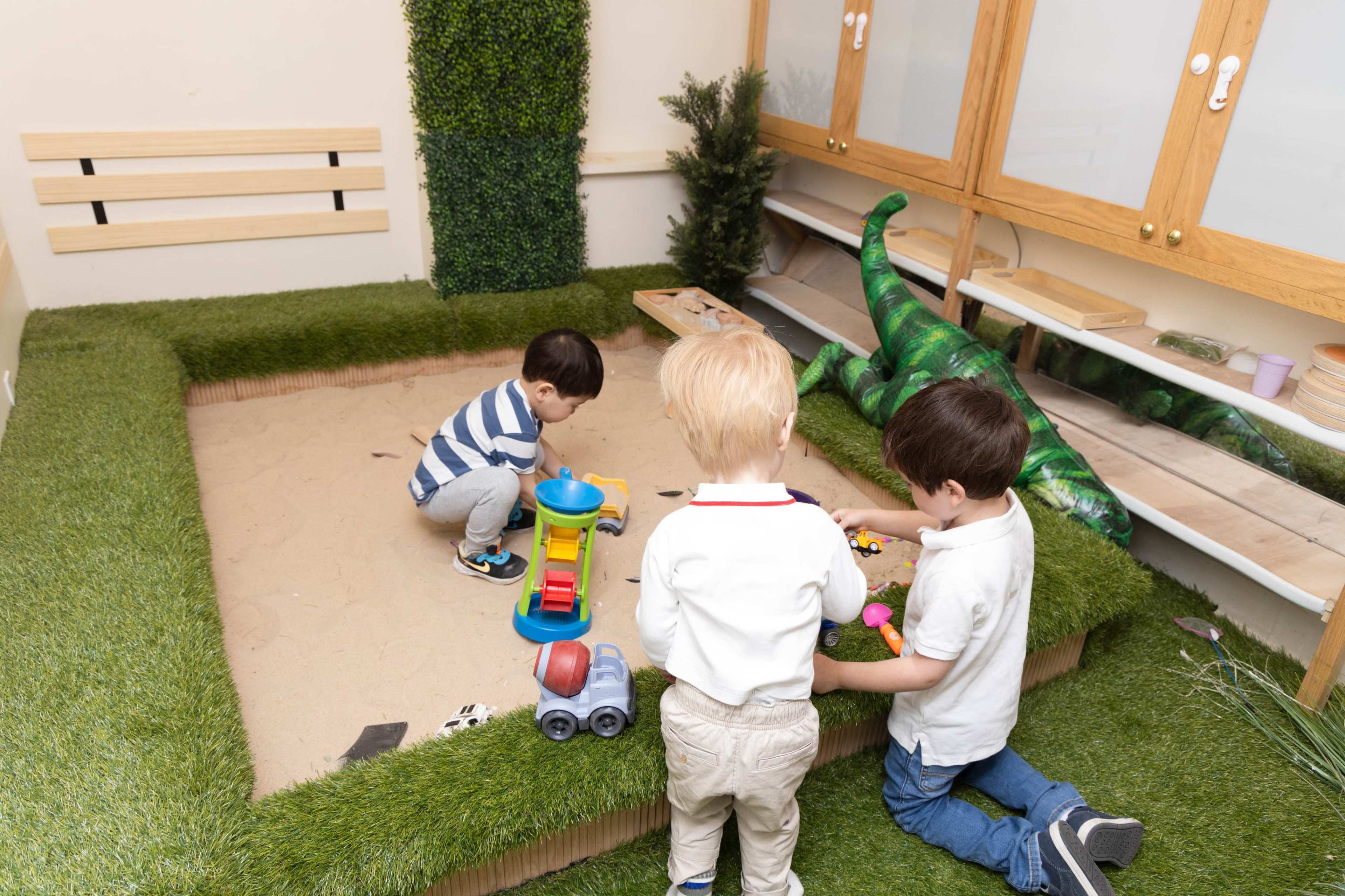 Three children play with toys in a sand play area surrounded by green grass-like turf.