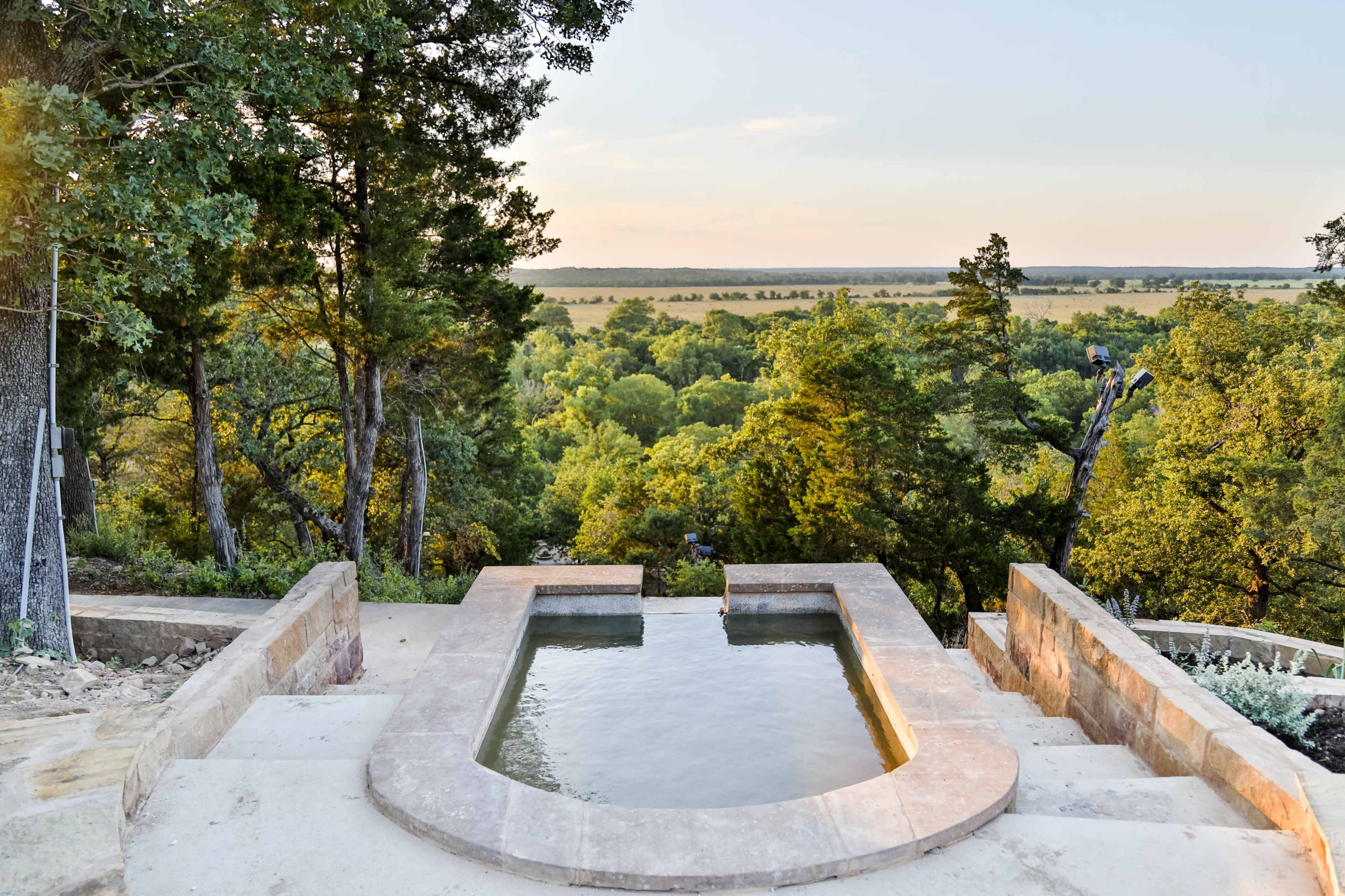 A rectangular, stone-lined pool is situated on a hillside, overlooking a lush green landscape with trees and rolling fields.