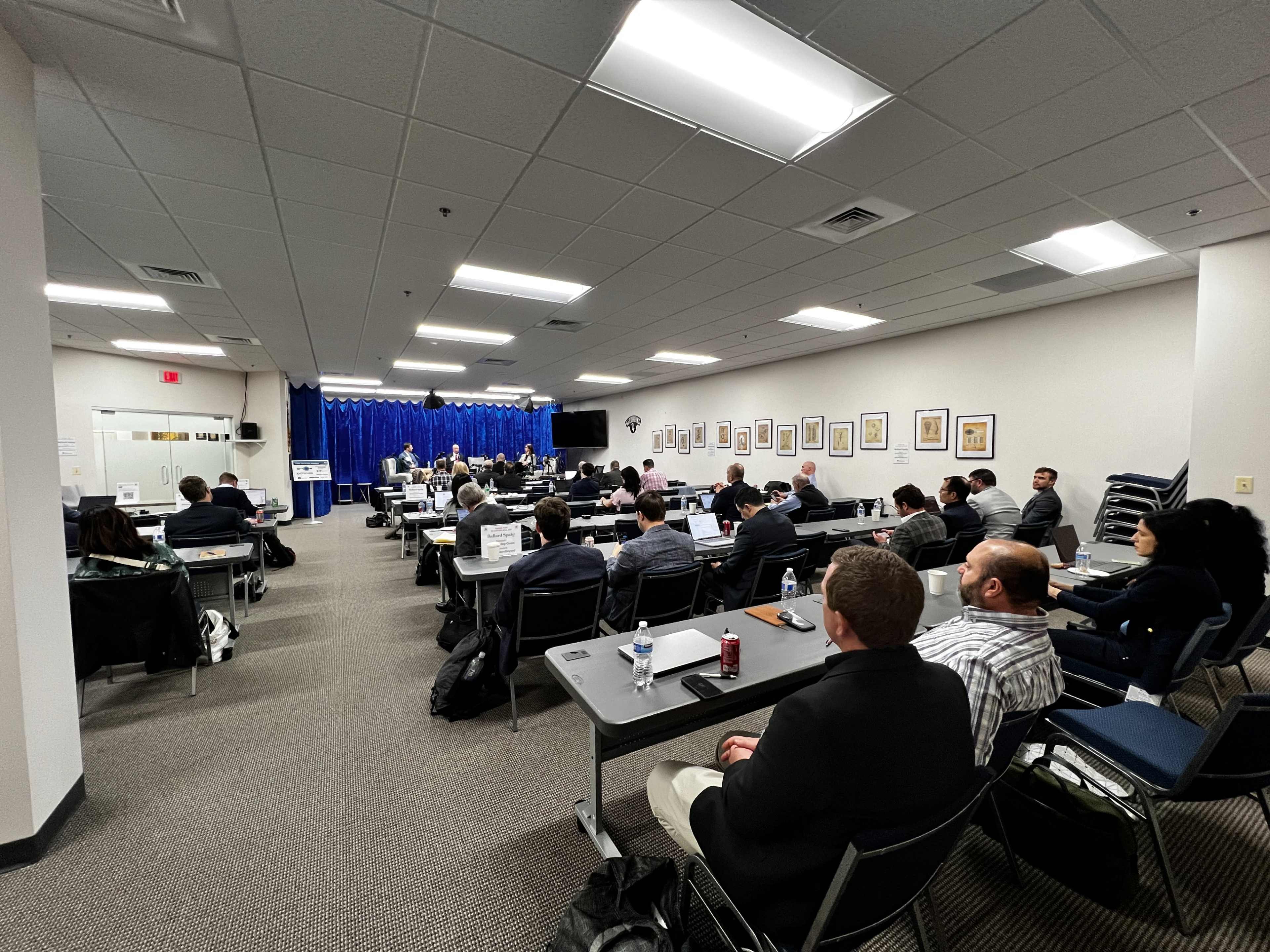 A group of attendees sits in a conference room facing a stage, where speakers are presenting in front of a blue curtain.