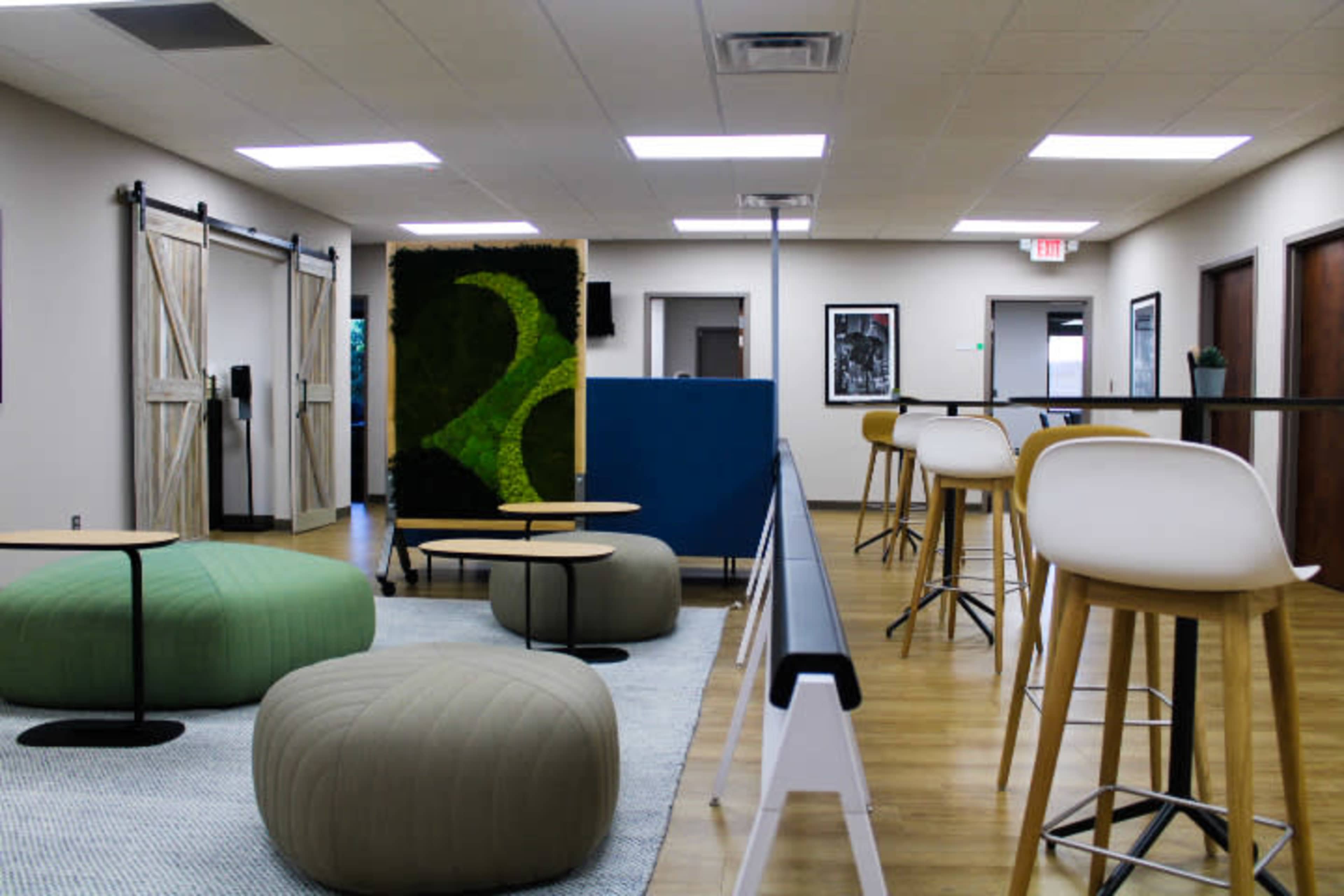 A modern office space with seating areas featuring round poufs and bar stools, surrounded by greenery and tasteful decor.