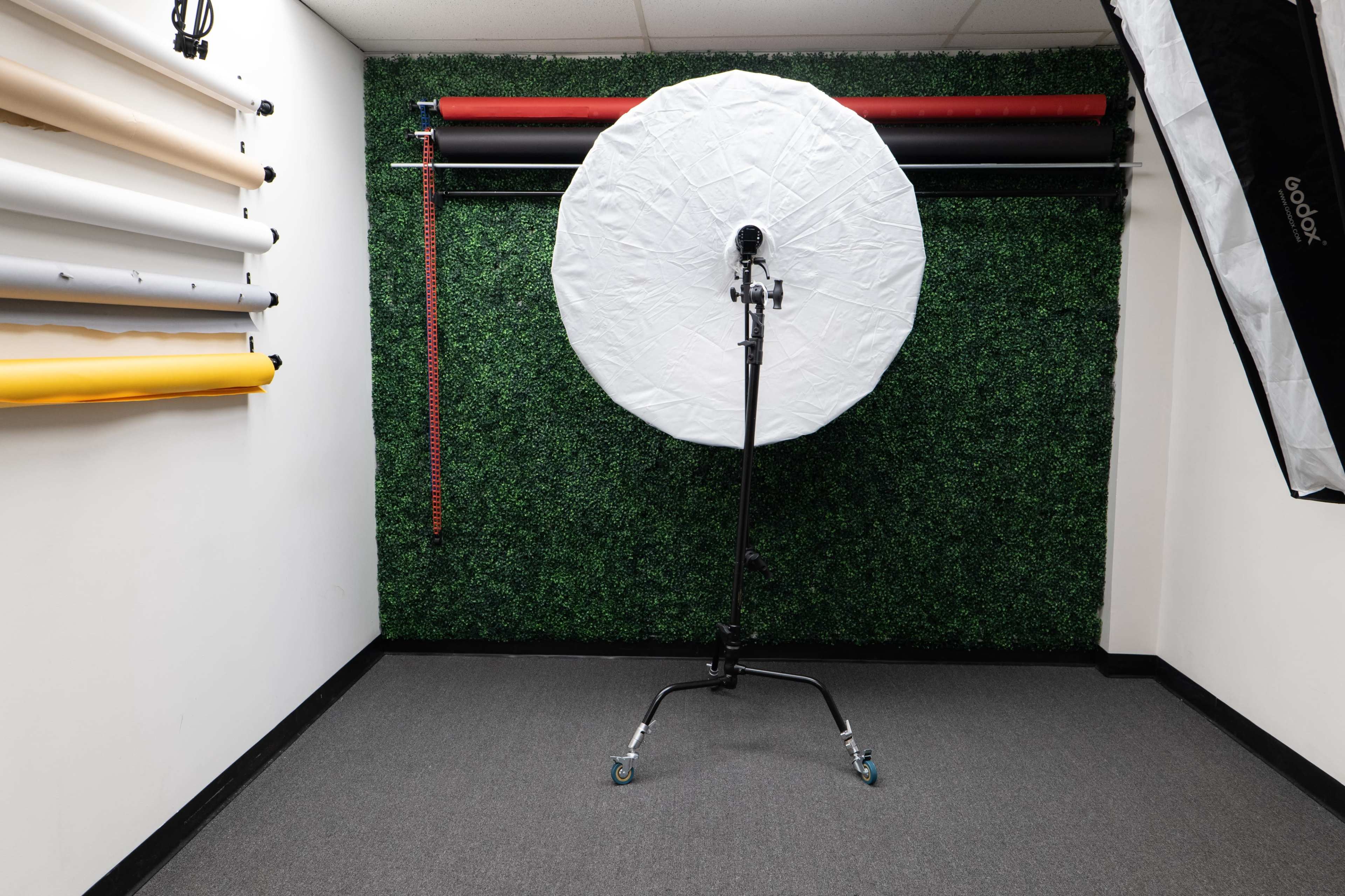 A photography studio with a round white umbrella softbox on a stand, set against a wall covered in artificial grass, with rolls of backdrops hanging on the side.