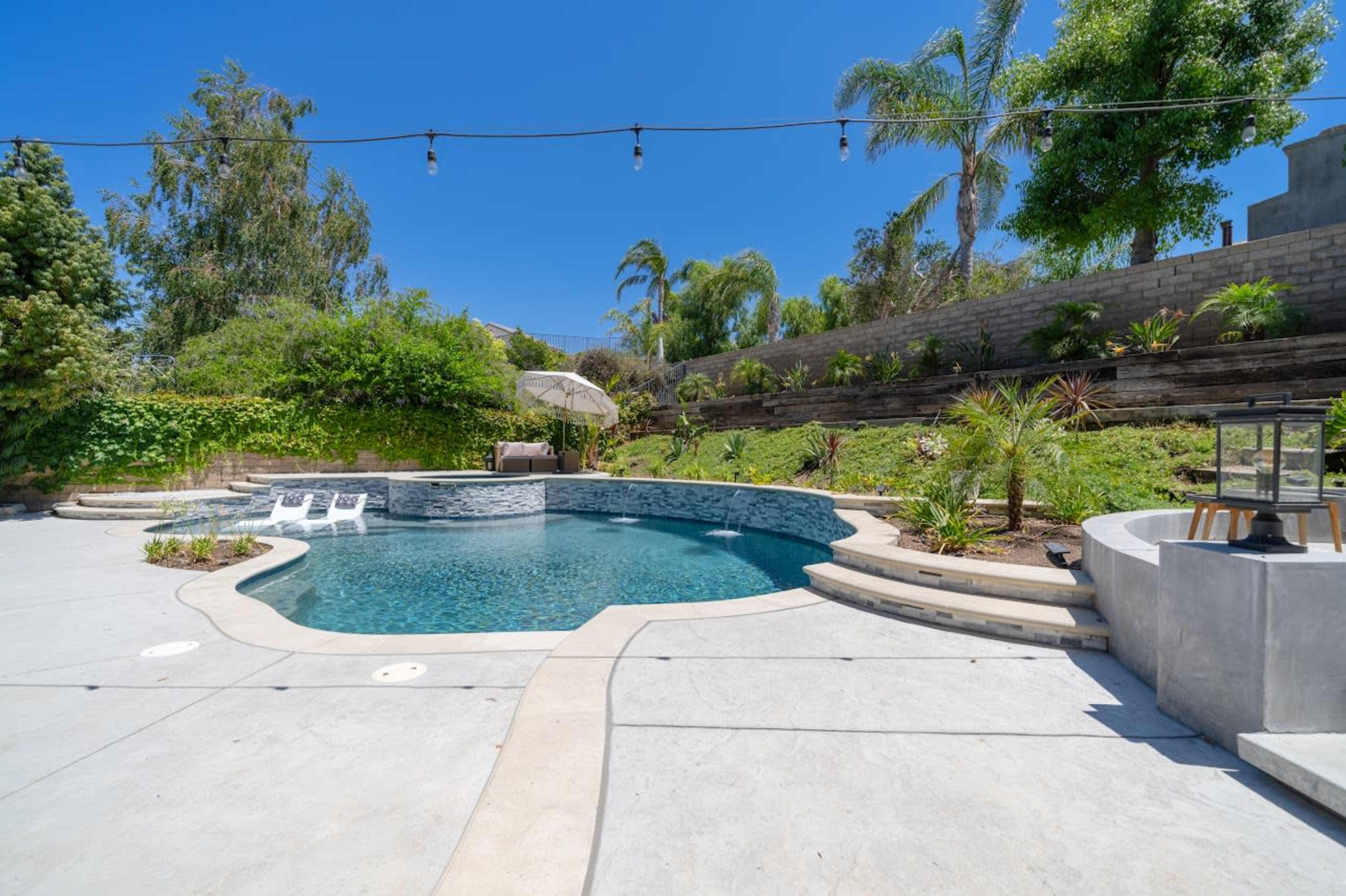 A backyard with a swimming pool surrounded by concrete, lounge chairs, and landscaping featuring palm trees and shrubs.