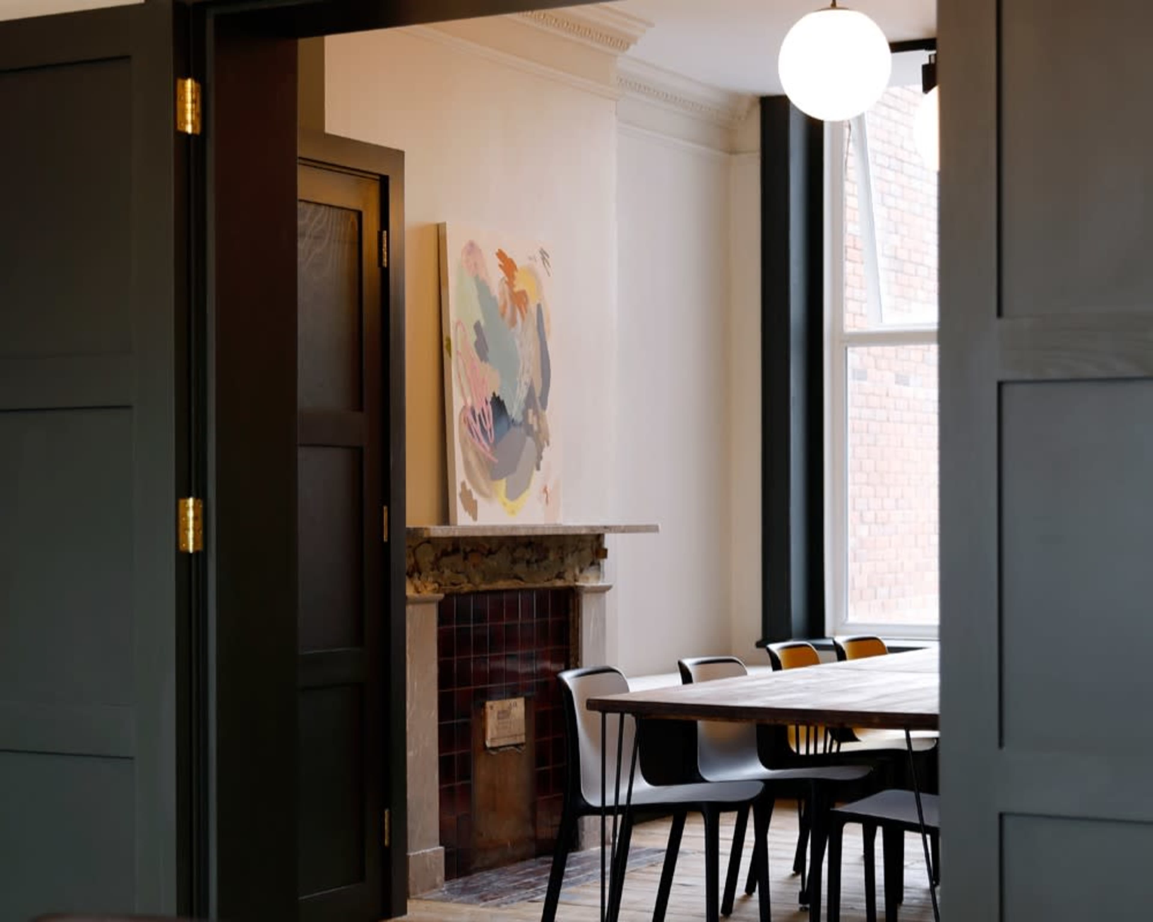 A dining area features a wooden table surrounded by several chairs, with an abstract painting on the wall and large windows letting in natural light.