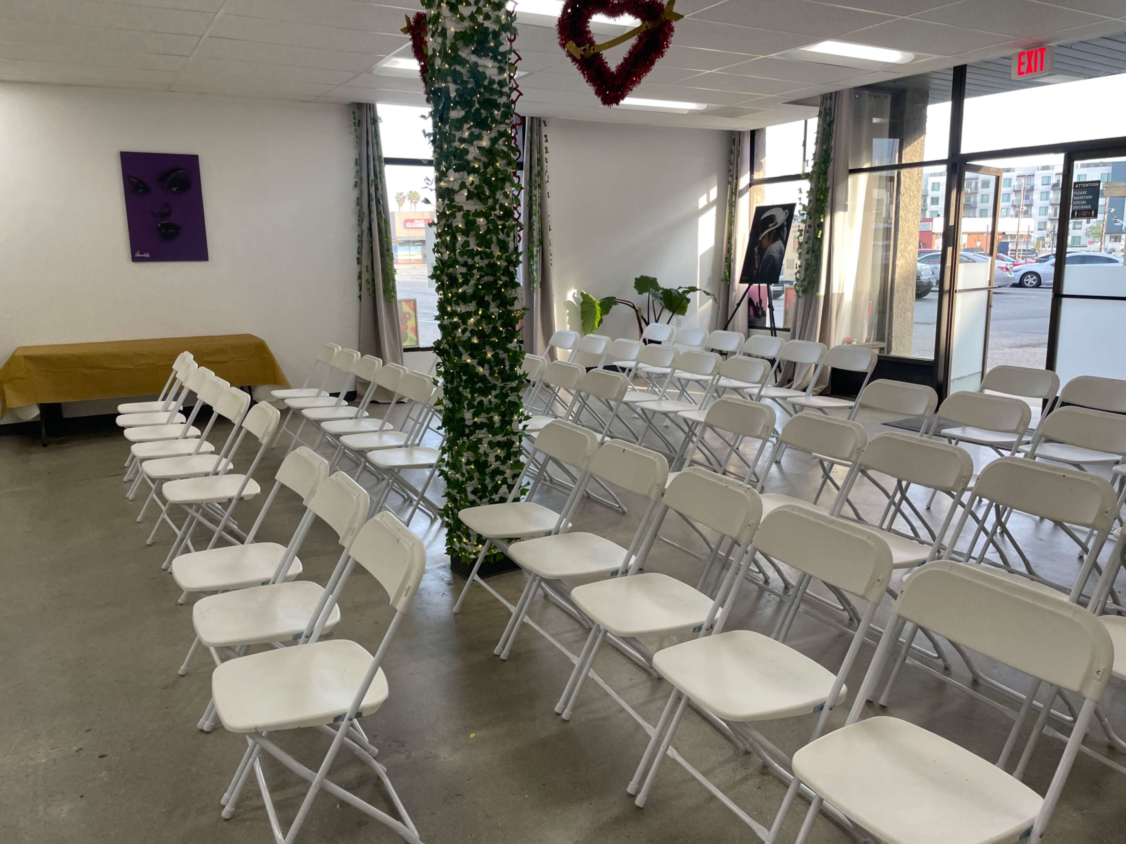 The image shows a venue set up for an event, featuring rows of white folding chairs arranged in two sections, with a decorative plant column and a purple artwork on the wall.