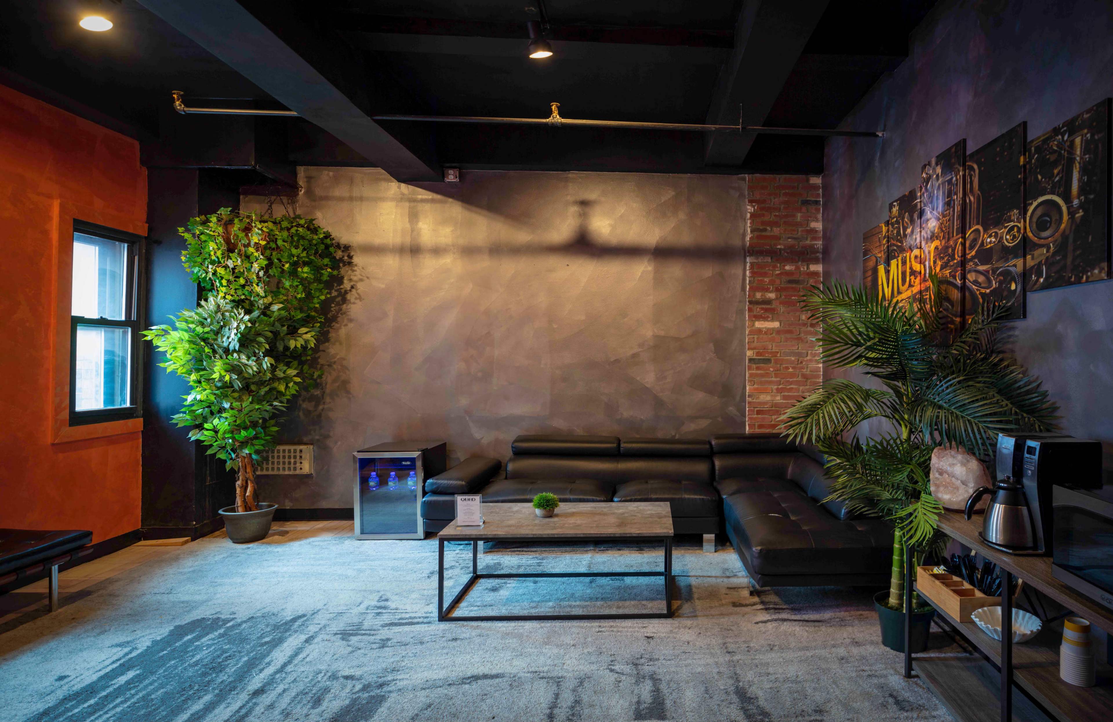 A modern lounge area featuring a black sectional sofa, a coffee table, a mini fridge, and decorative plants against a backdrop of textured walls and artwork.