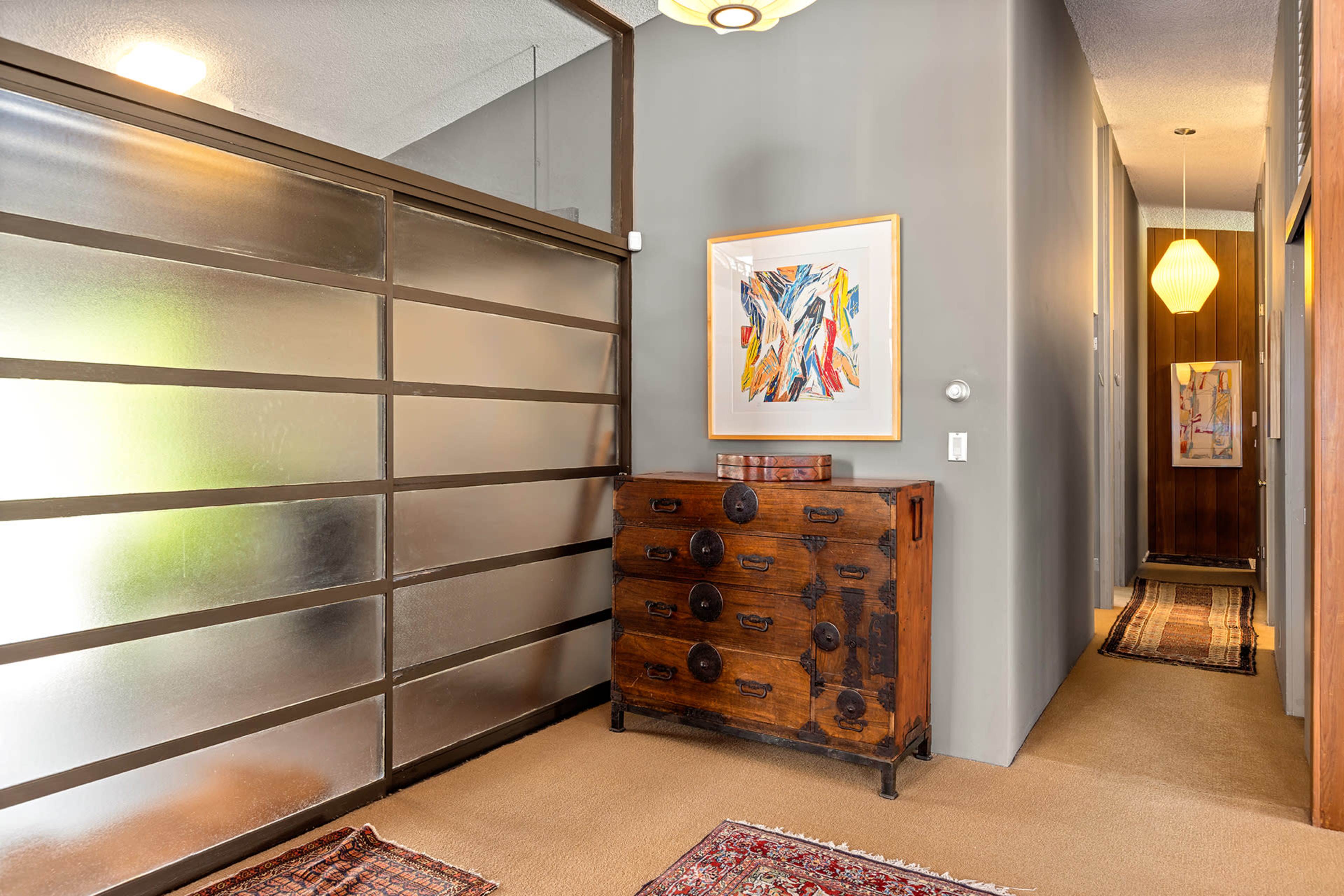 The image shows a hallway with a frosted glass partition, a wooden chest of drawers, and artwork on the wall, leading to a door at the far end.