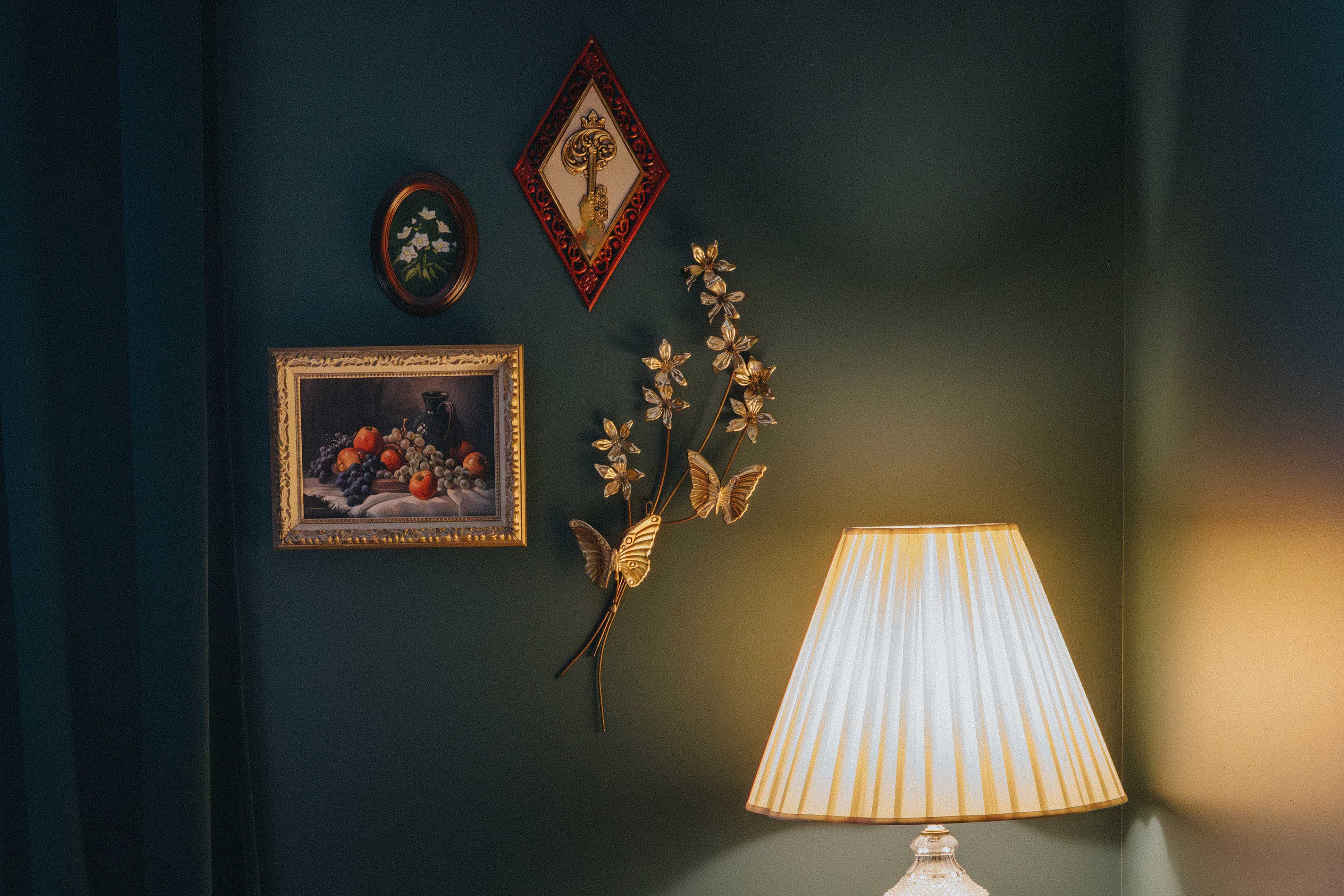 A lamp with a pleated shade stands beside a dark green wall adorned with framed artwork and decorative elements including flowers and butterflies.