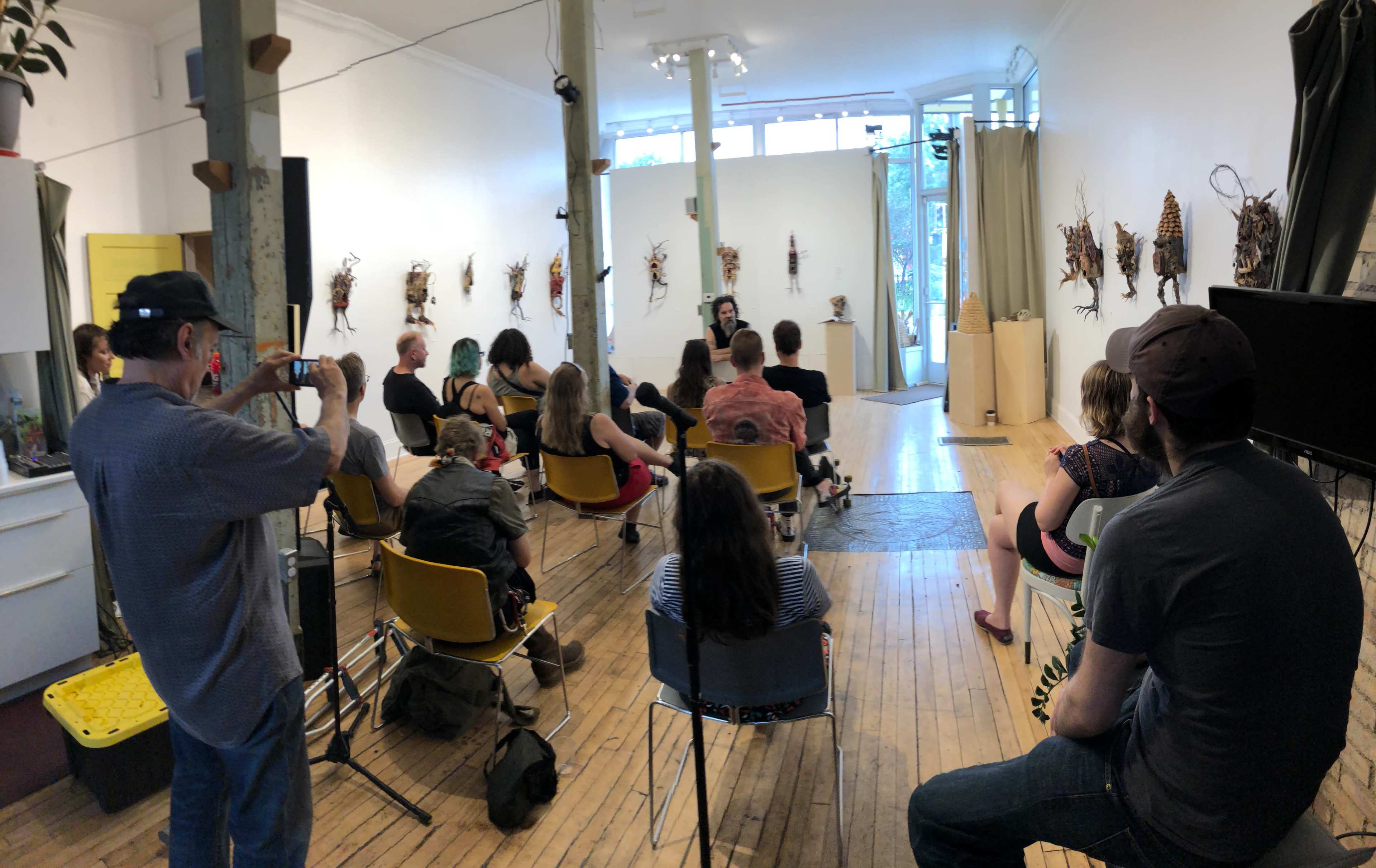 A group of people sits in chairs facing a speaker in an art gallery adorned with various natural displays on the walls.
