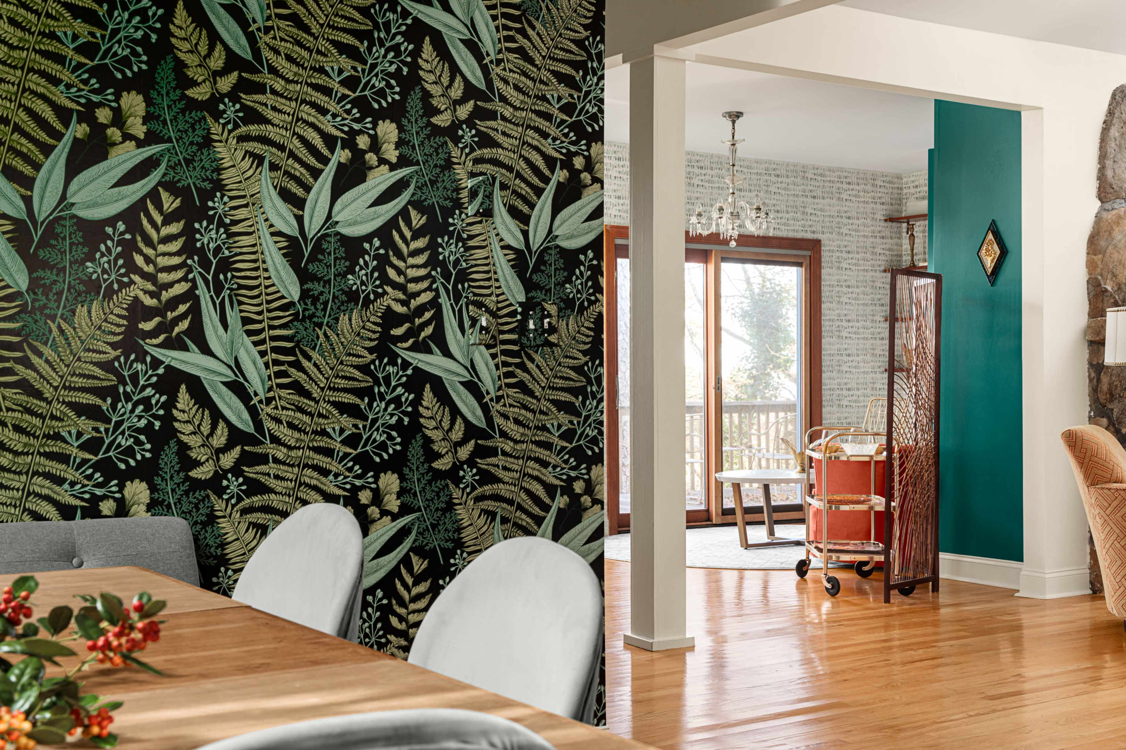 A dining area with a botanical wallpaper, an open doorway leading to a living space with a green wall and a wooden cart.