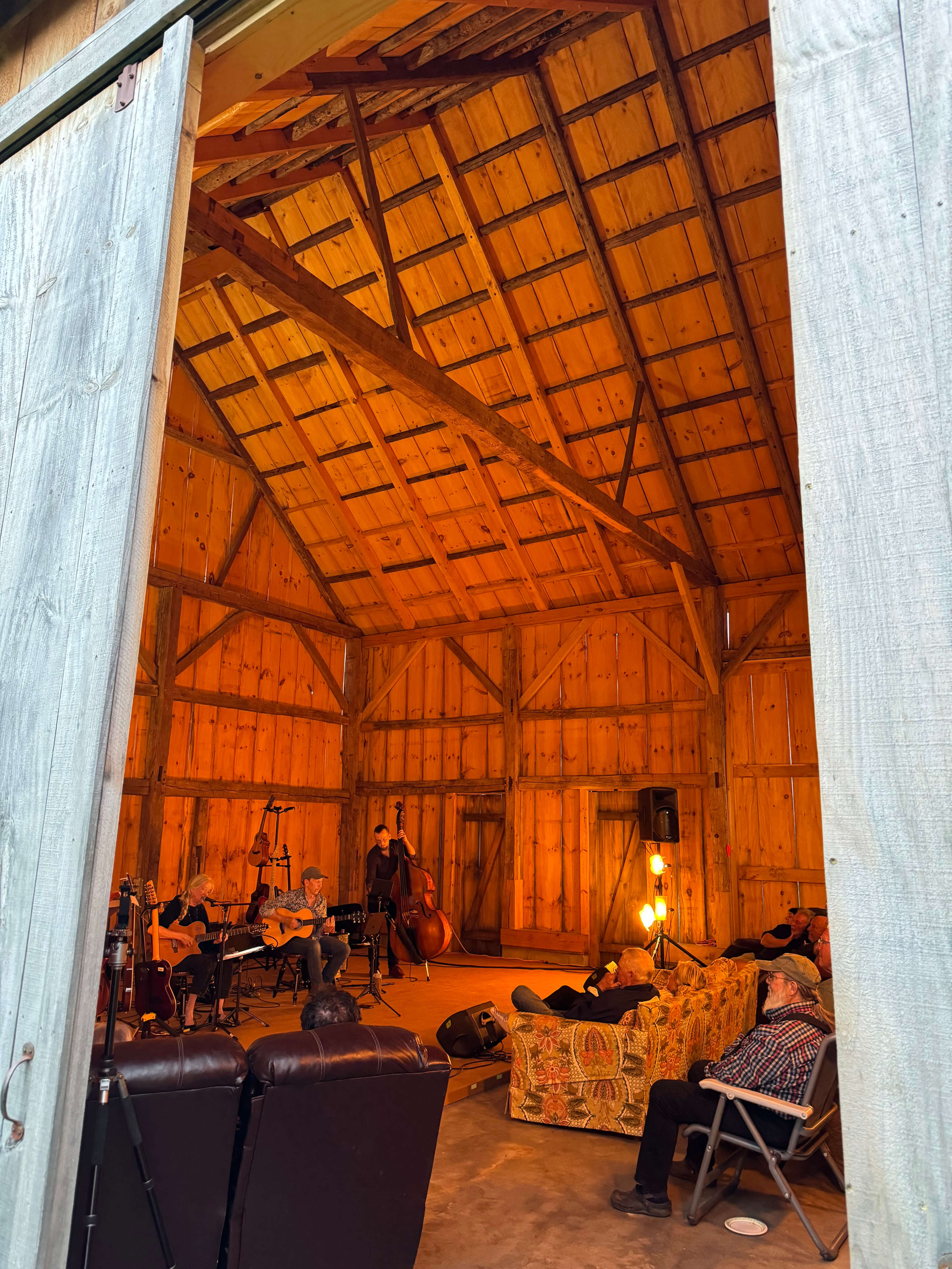 A group of musicians performs inside a wooden barn with warm lighting, while several people relax on couches in the foreground.