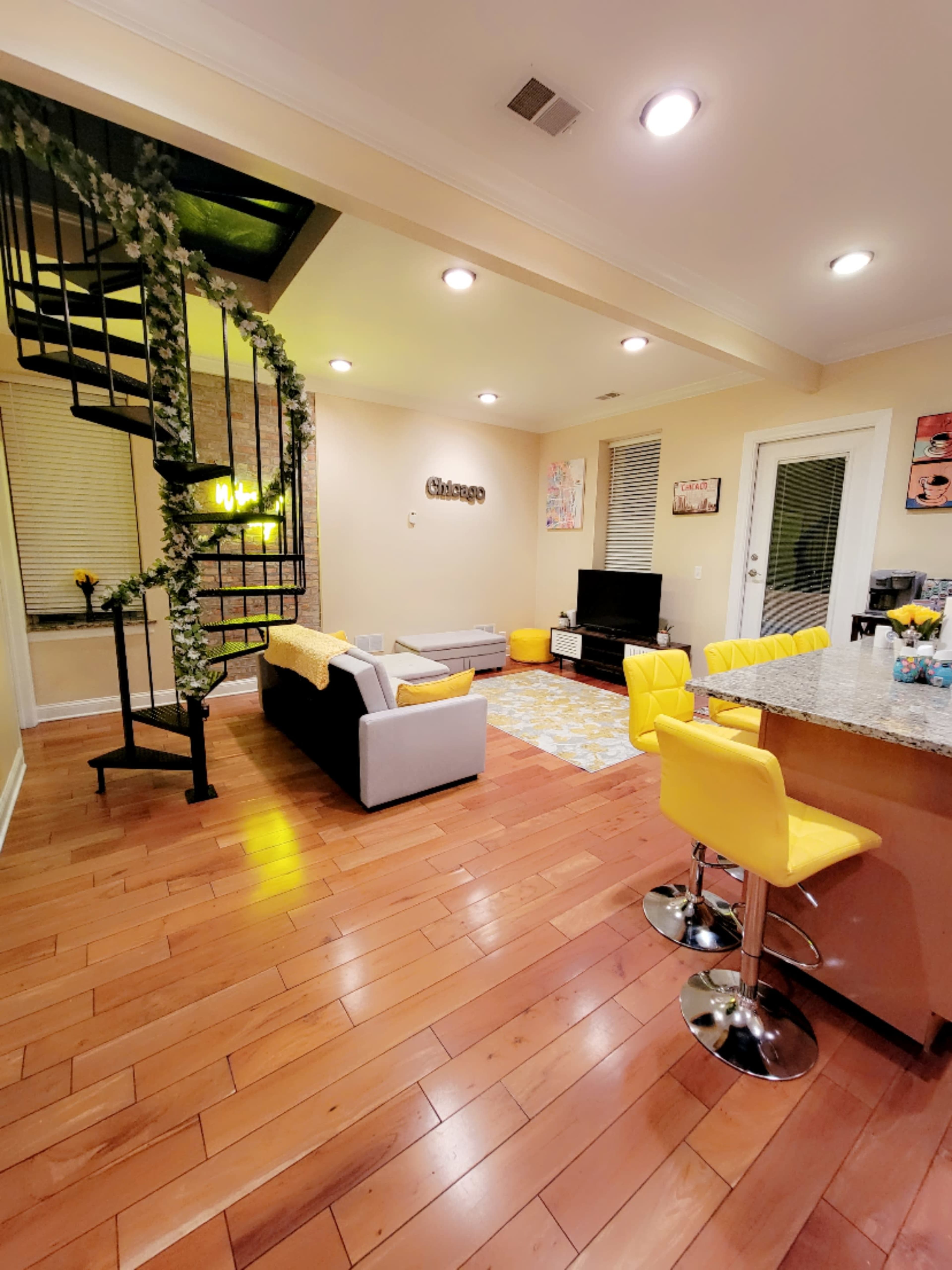 A modern living room and kitchen area with wooden flooring, a spiral staircase, yellow bar stools, and a gray sofa.