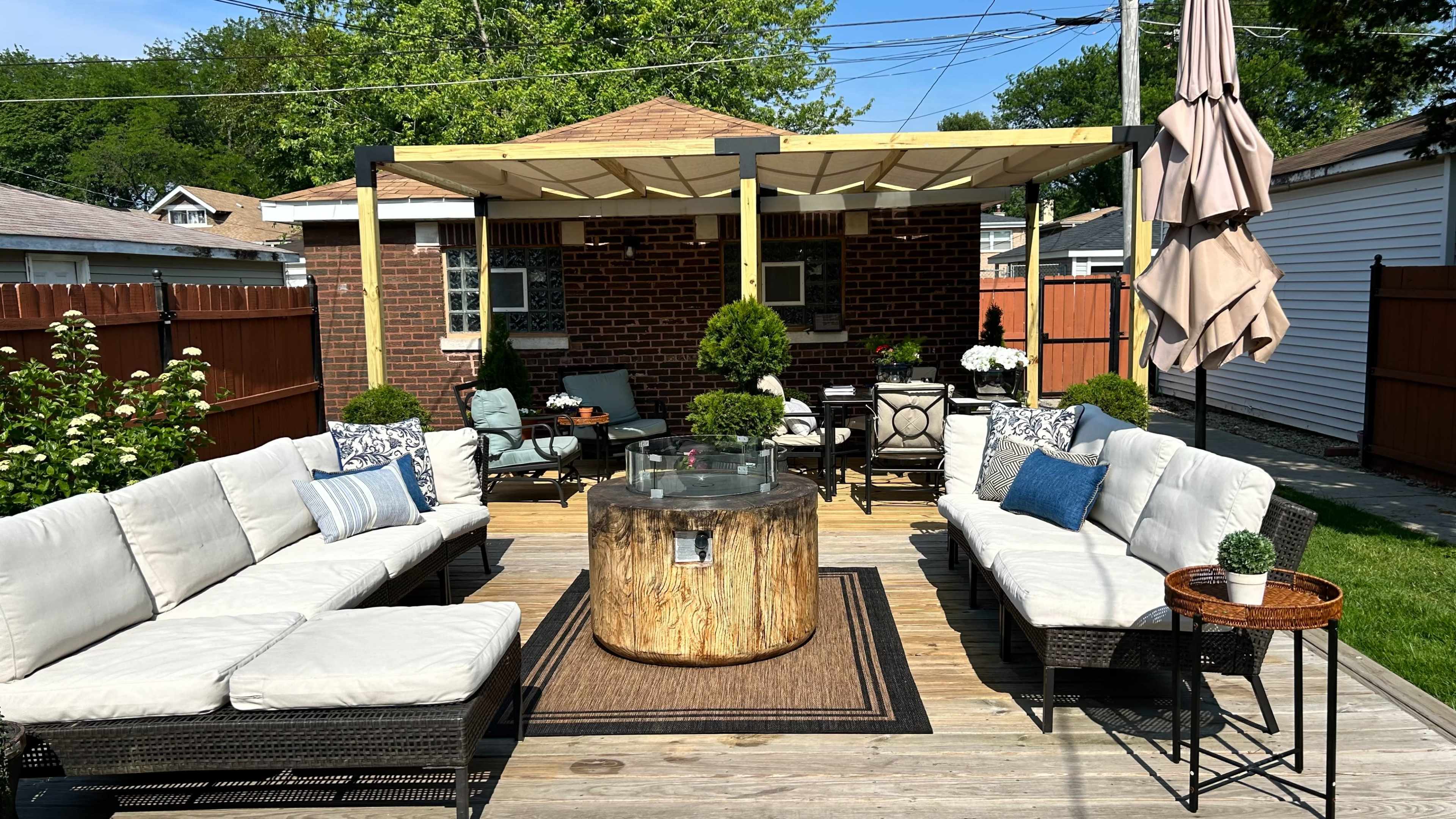 The image shows a well-furnished outdoor patio with a circular wooden table, surrounded by sectional sofas and various potted plants.