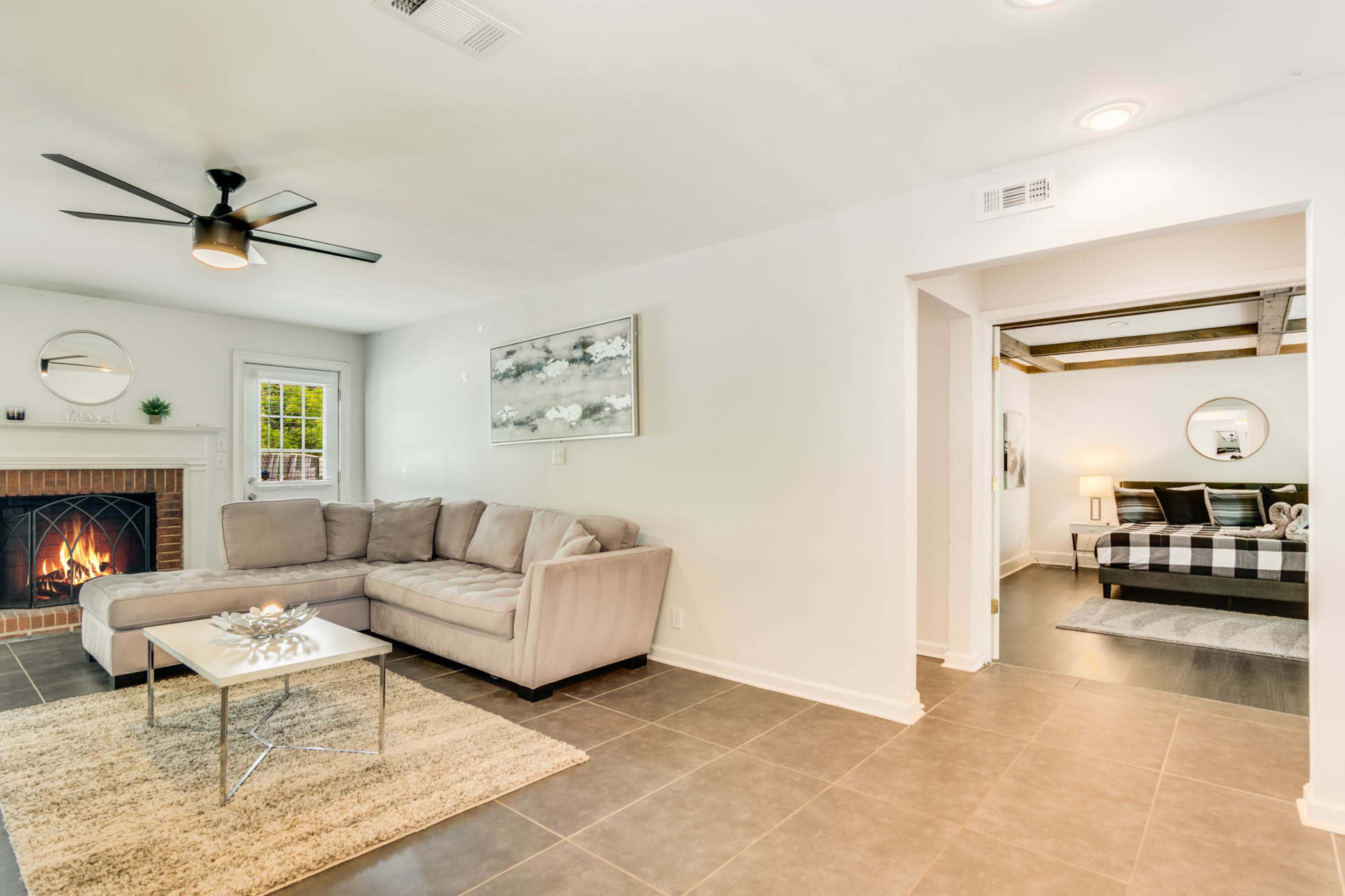The image shows a modern living room with a sectional sofa, a coffee table, and a fireplace, connected to a bedroom area in the background.