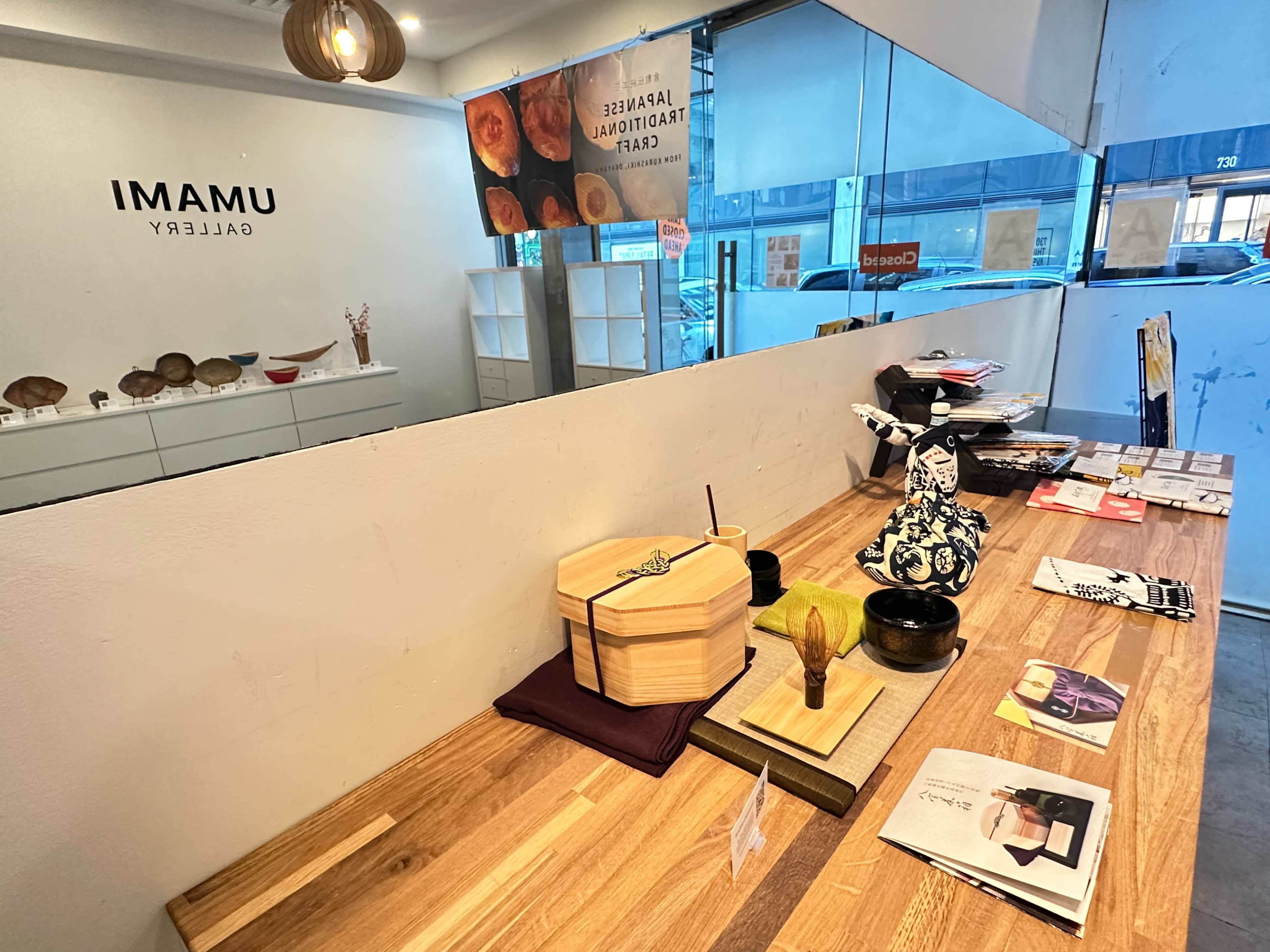 A sleek wooden table in a gallery featuring a display of art materials, utensils, and a decorative box, with a modern white wall and large windows in the background.