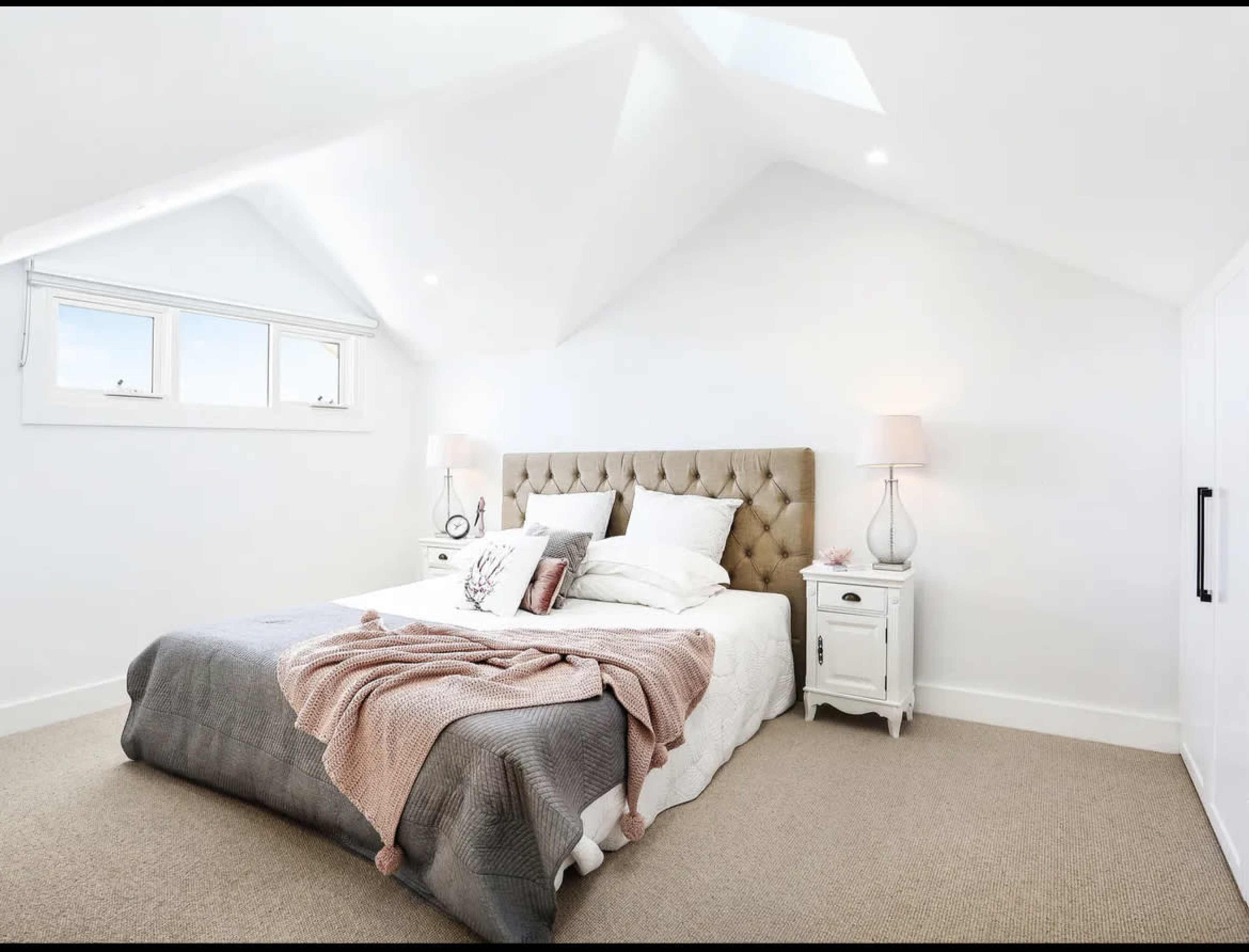 A well-lit, minimalist bedroom features a beige tufted headboard, white bedding with decorative pillows, and two bedside tables with lamps.