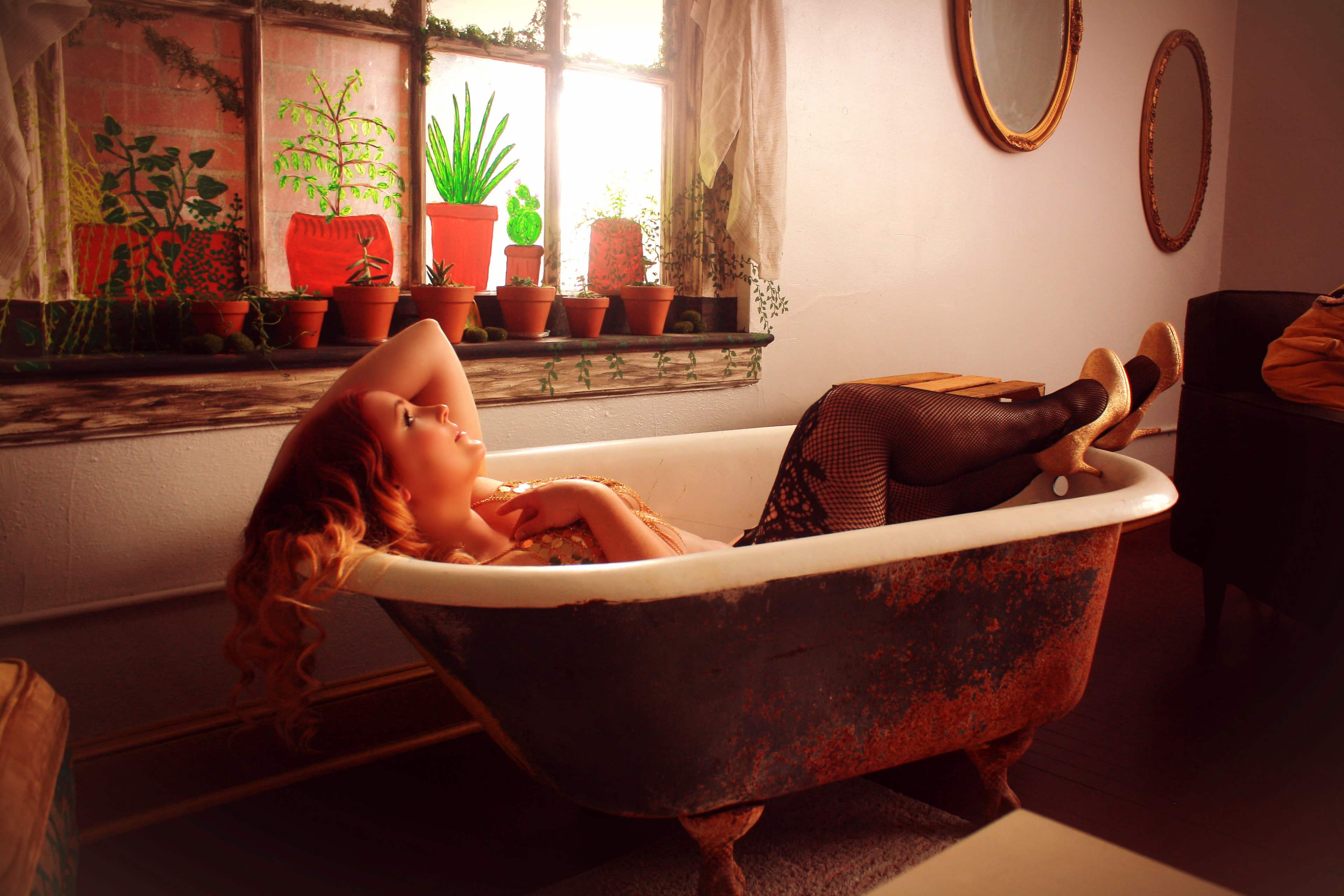A woman with long hair lounges in a vintage bathtub surrounded by potted plants and natural light.