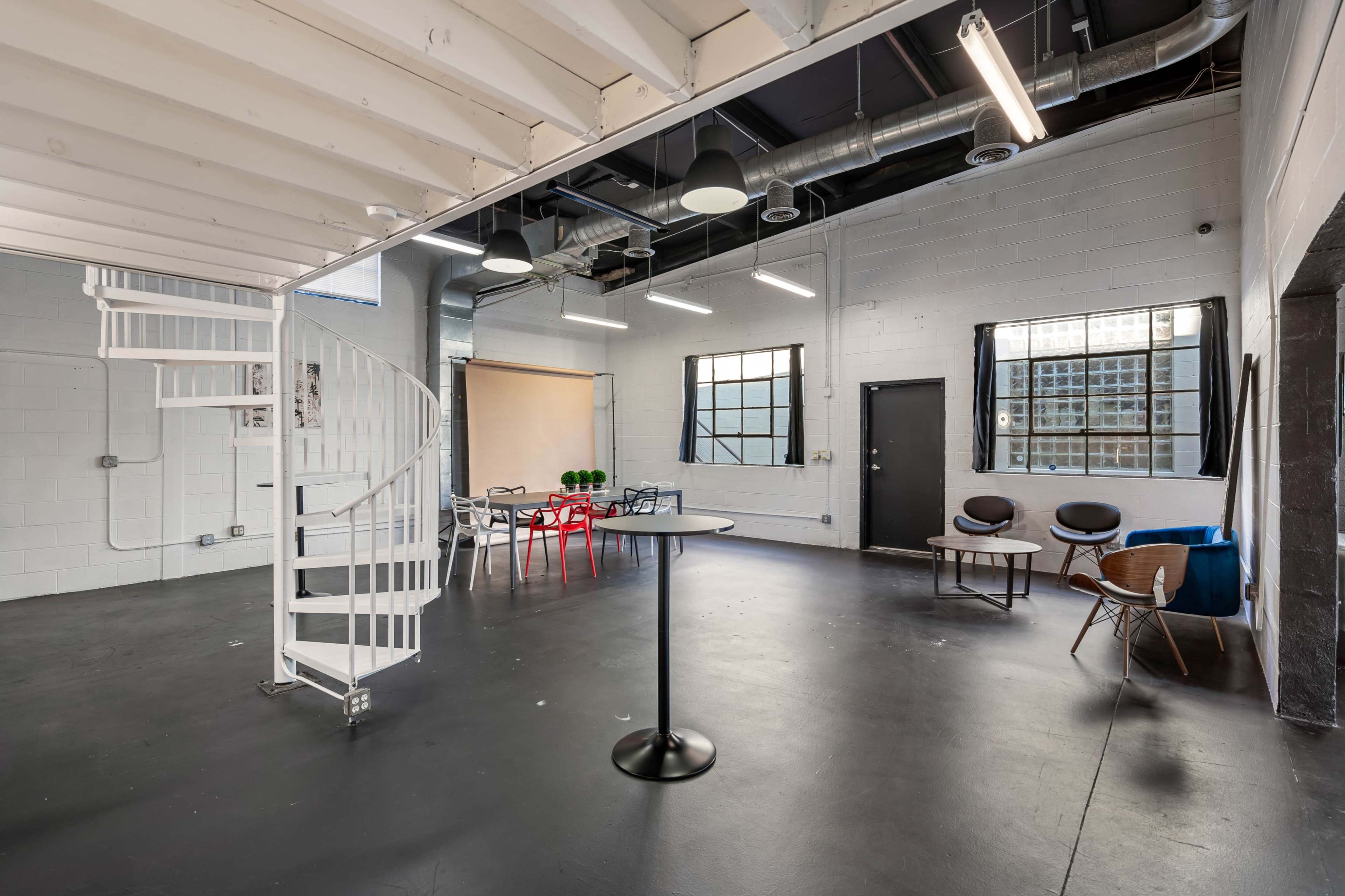 A spacious, industrial-style interior with a spiral staircase, black flooring, large windows, and a mixture of seating and dining areas.