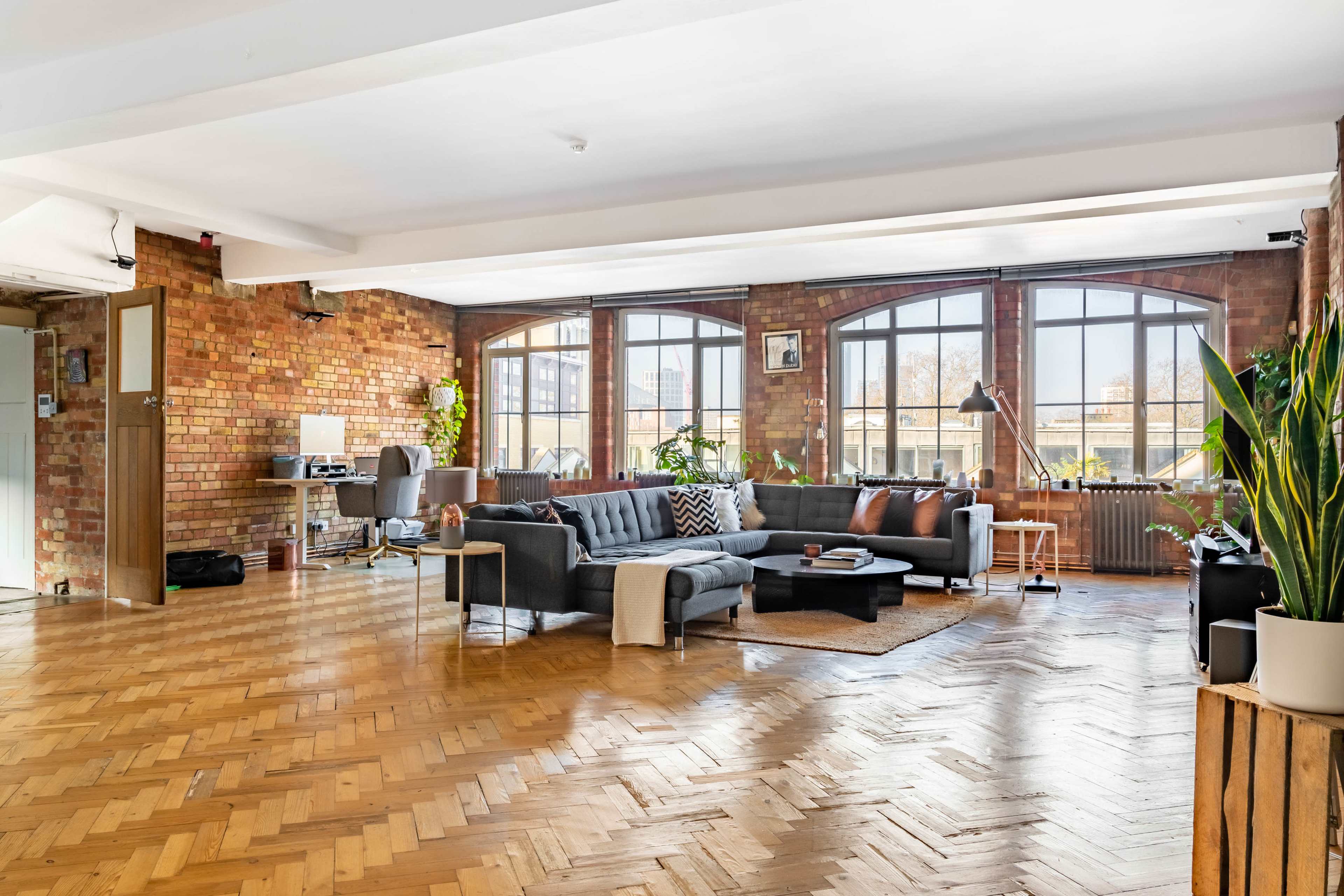 A spacious, modern living area with a gray sectional couch, wooden floors, and large windows allowing natural light to fill the room.