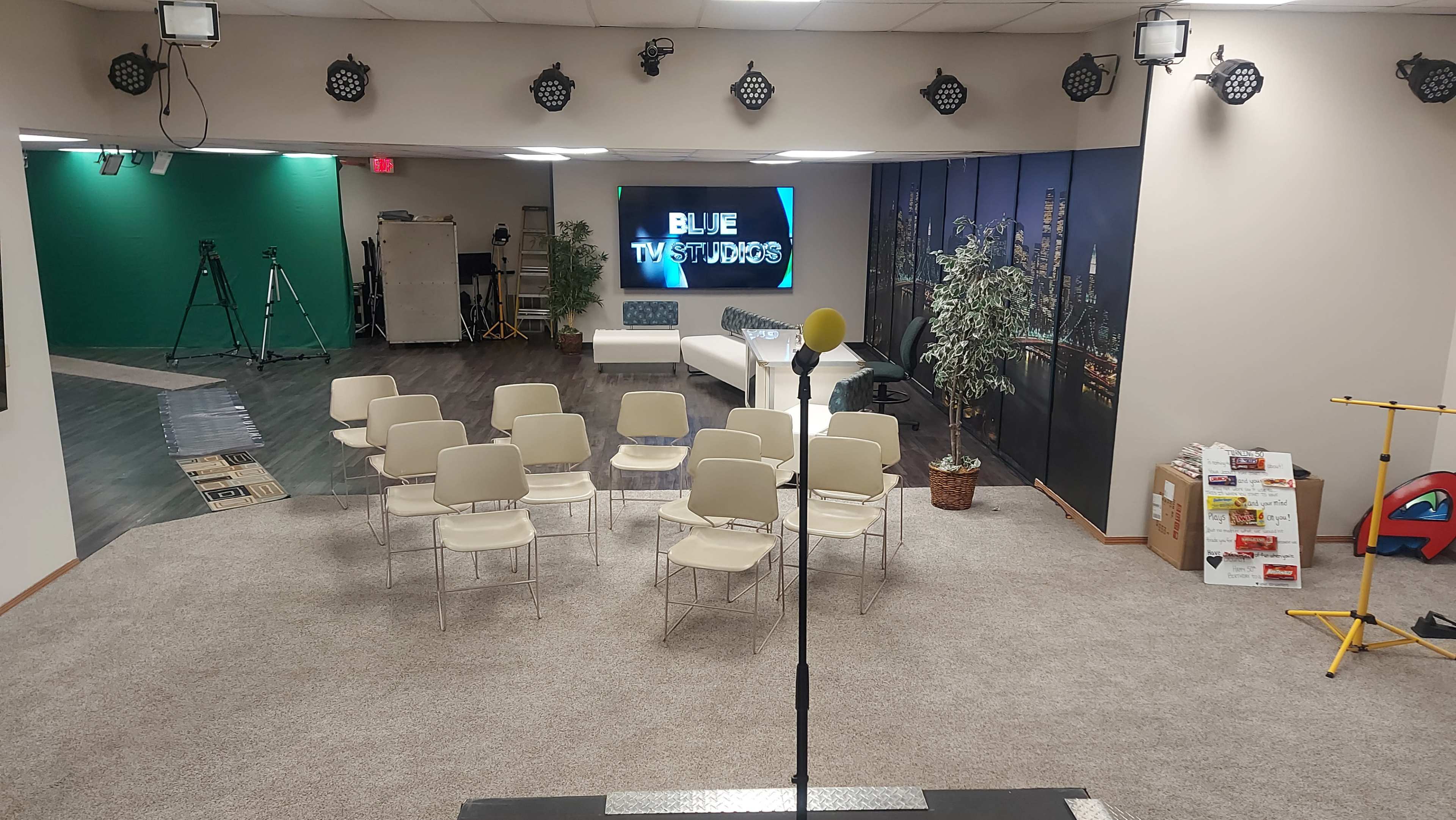 An empty studio space with rows of chairs facing a microphone on a stage, flanked by green and black backdrop setups.