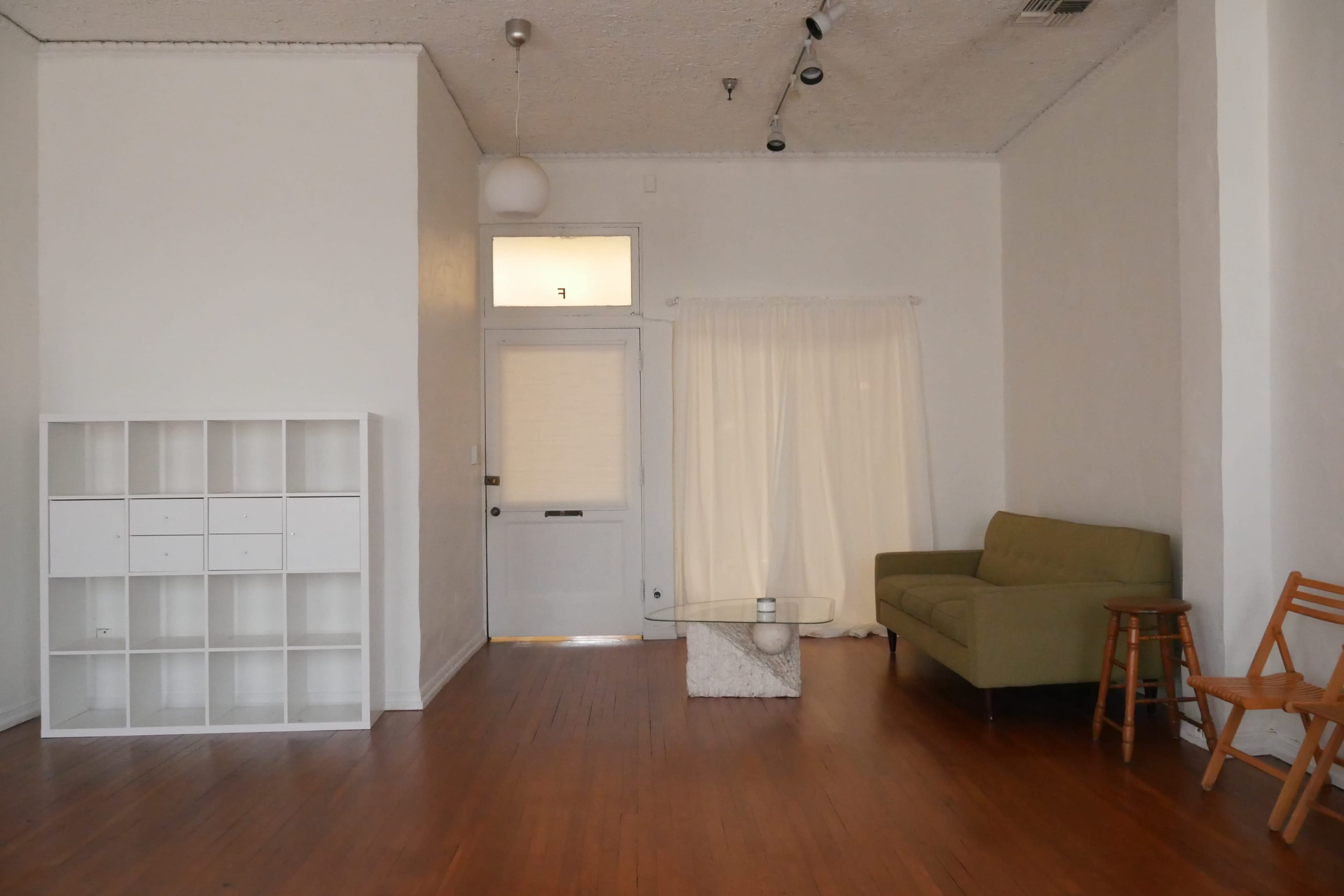 The image shows a minimalist living space with a green sofa, a glass coffee table, a wooden chair, and a white shelving unit against a wall.