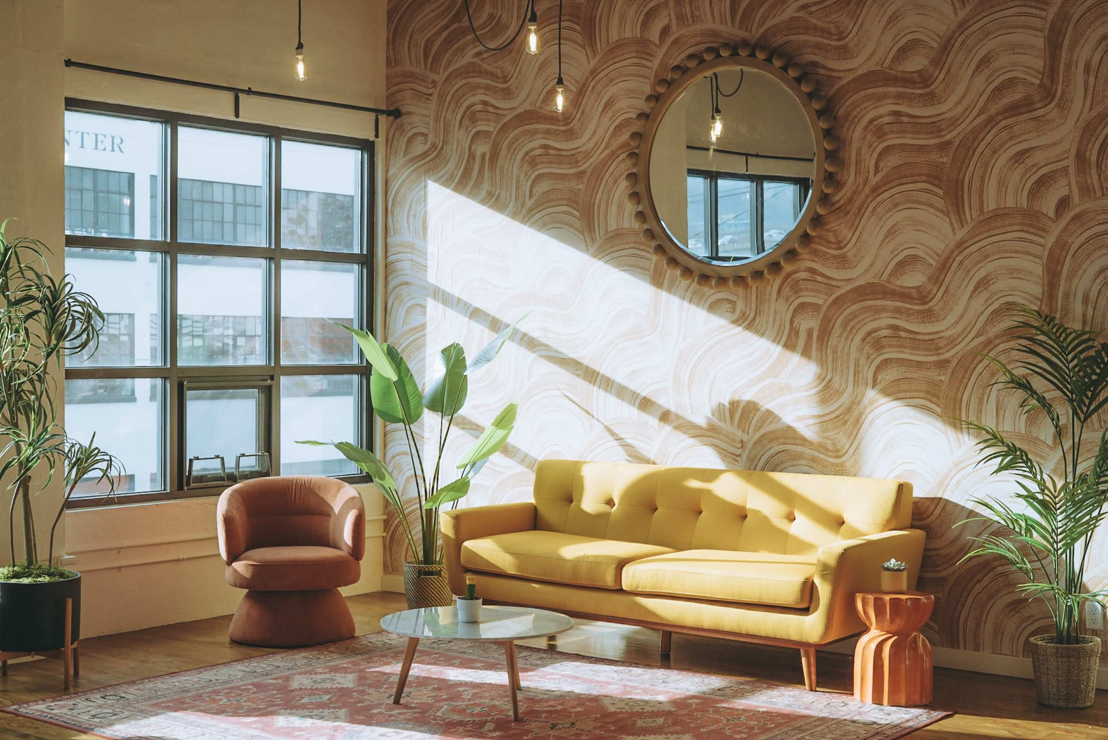 A cozy living room with a yellow couch, a round mirror, and a coffee table, illuminated by sunlight streaming through a large window.