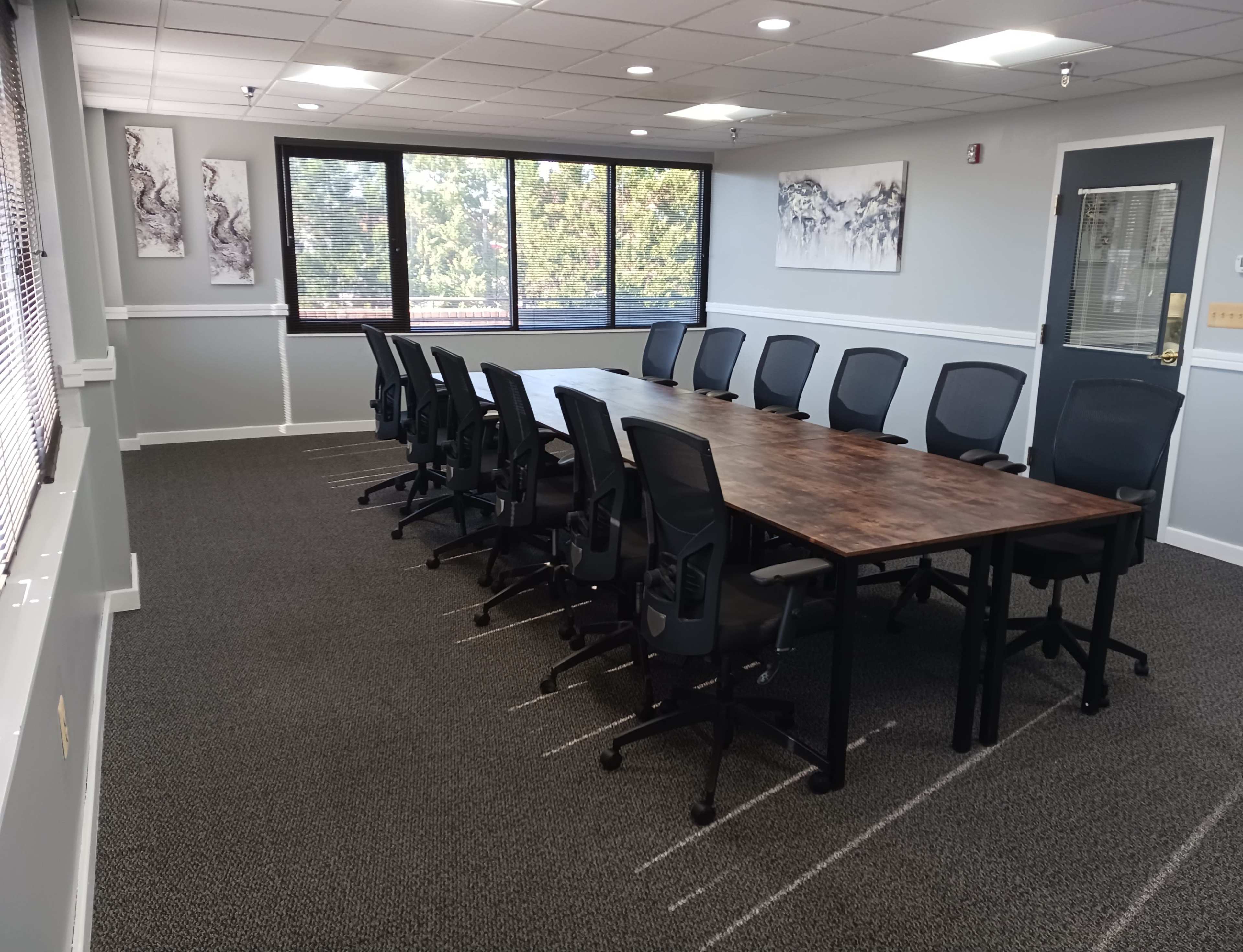 A conference room features a long wooden table surrounded by ergonomic black chairs, with large windows offering a view of greenery outside.