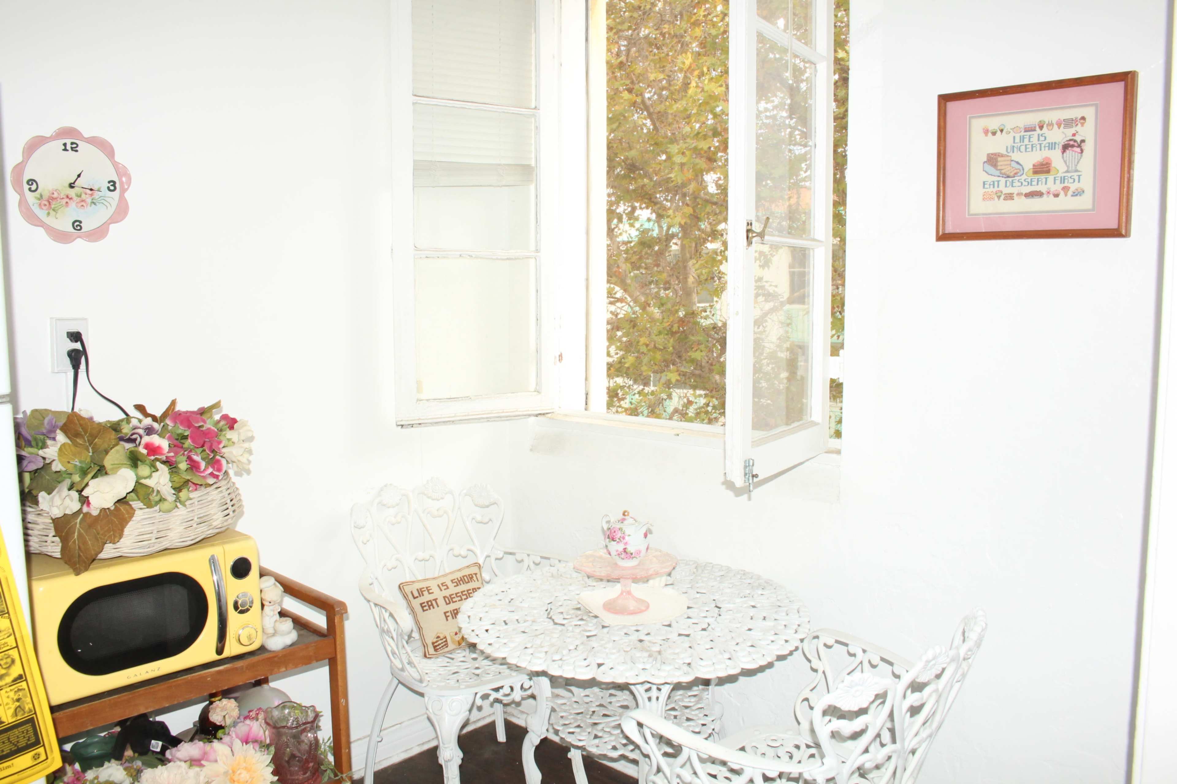 A small corner dining area features a round table with white wrought-iron chairs, a yellow microwave, and a window that opens to a view of trees.
