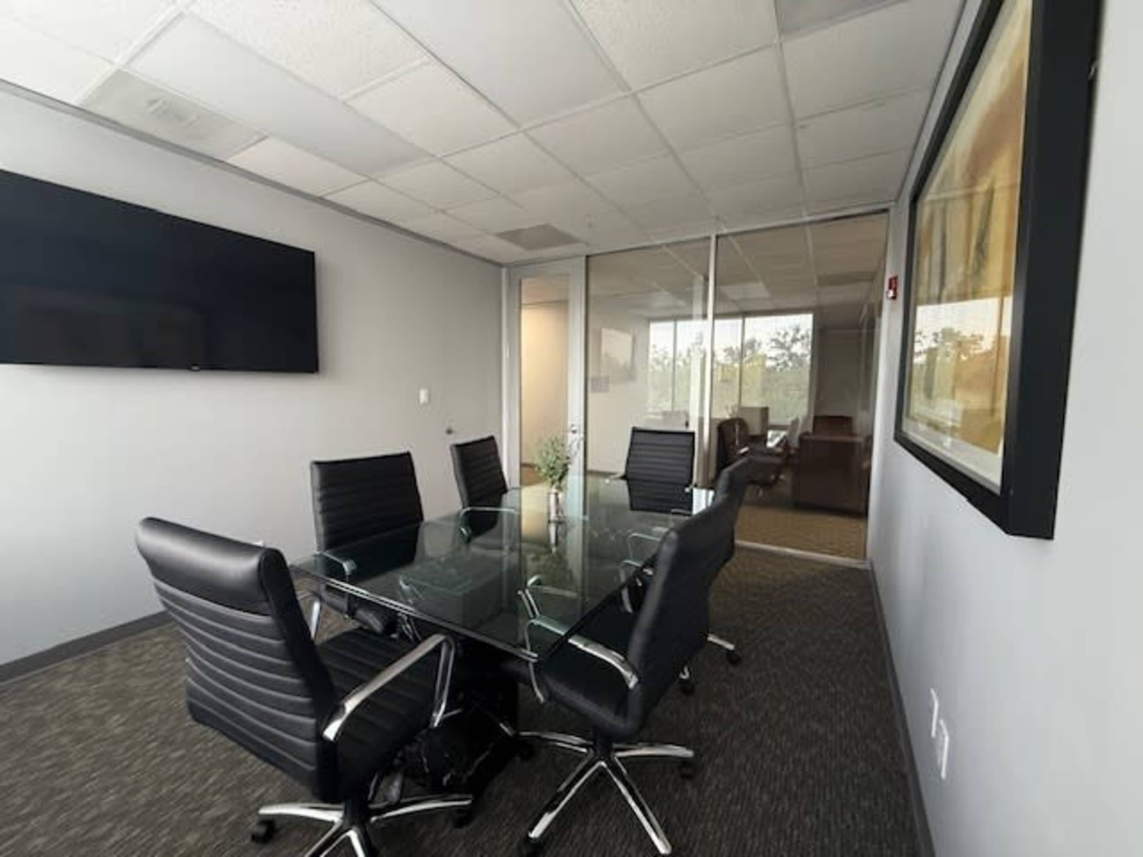 The image shows a modern conference room with a glass table surrounded by black office chairs, featuring a wall-mounted TV and a glass-walled office in the background.