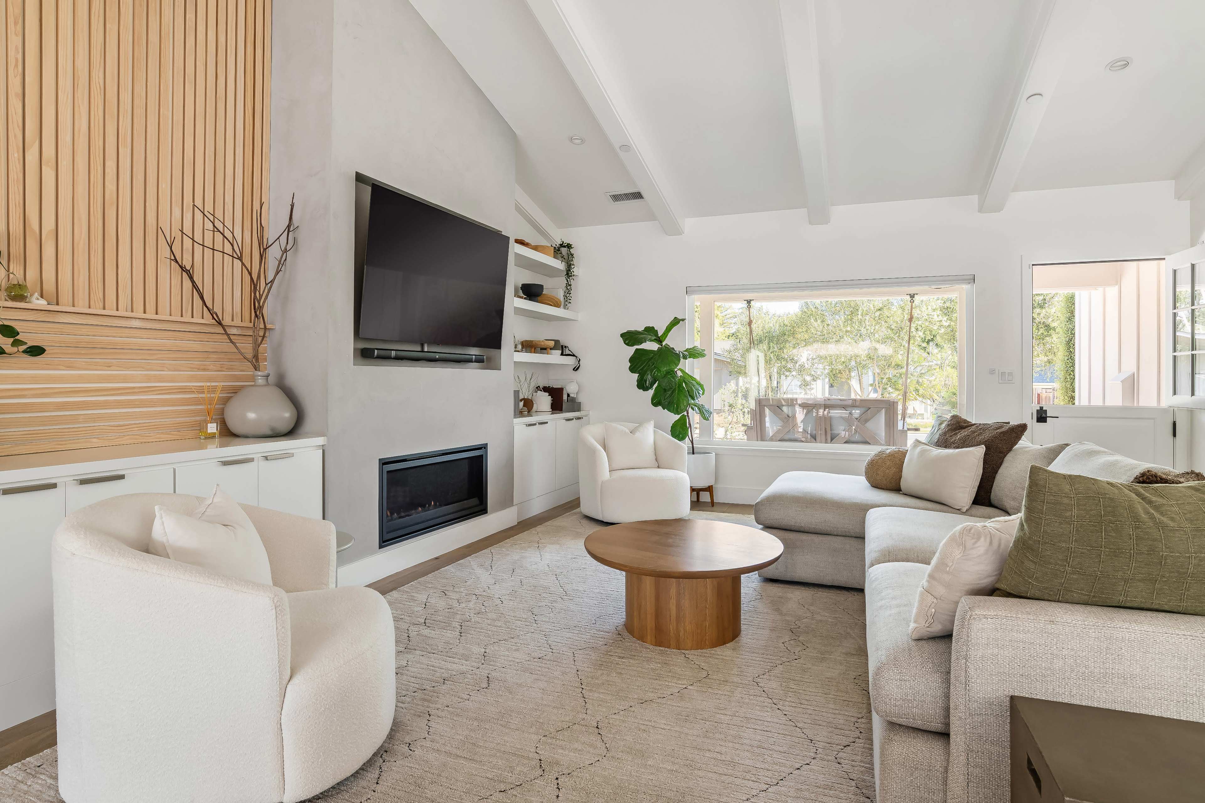 The image shows a modern living room featuring a sectional sofa, a round wooden coffee table, and a large wall-mounted television, with natural light streaming in through sliding glass doors.