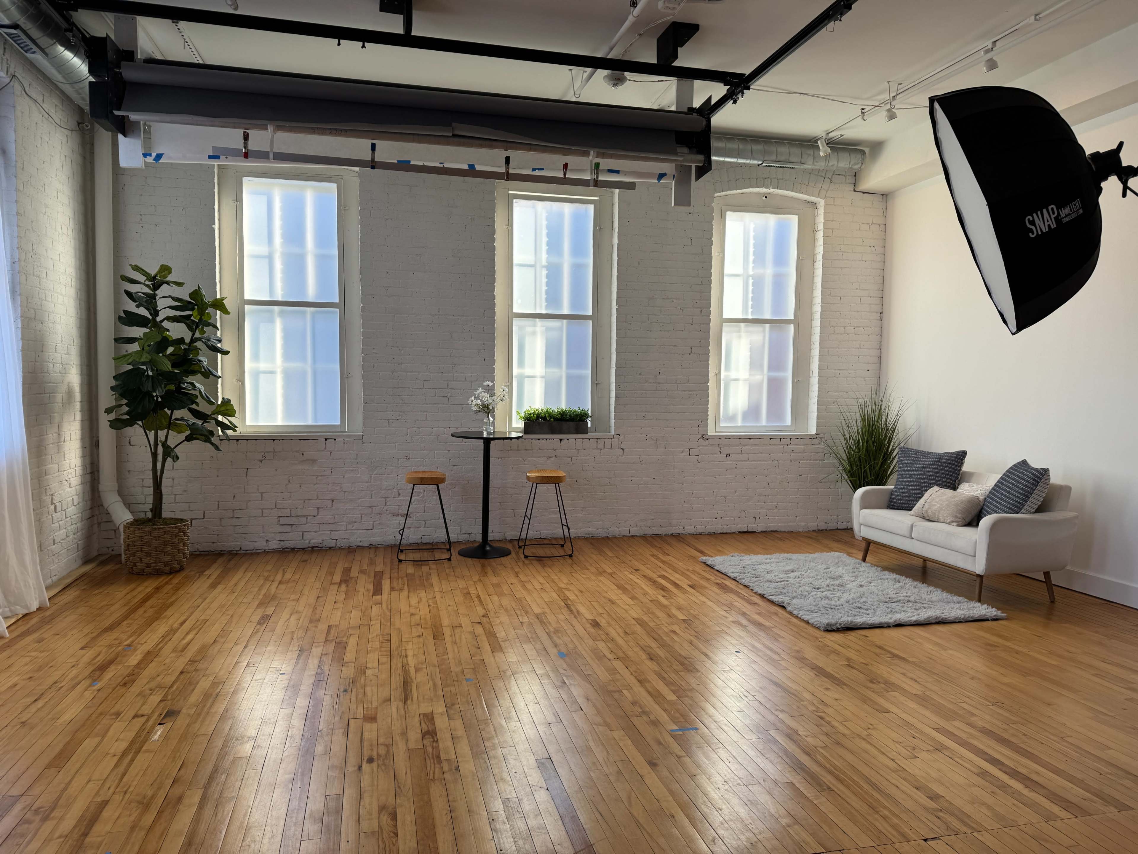 A bright, uncluttered studio space features two large windows, a small table with two stools, a sofa, and decorative plants on a wooden floor.