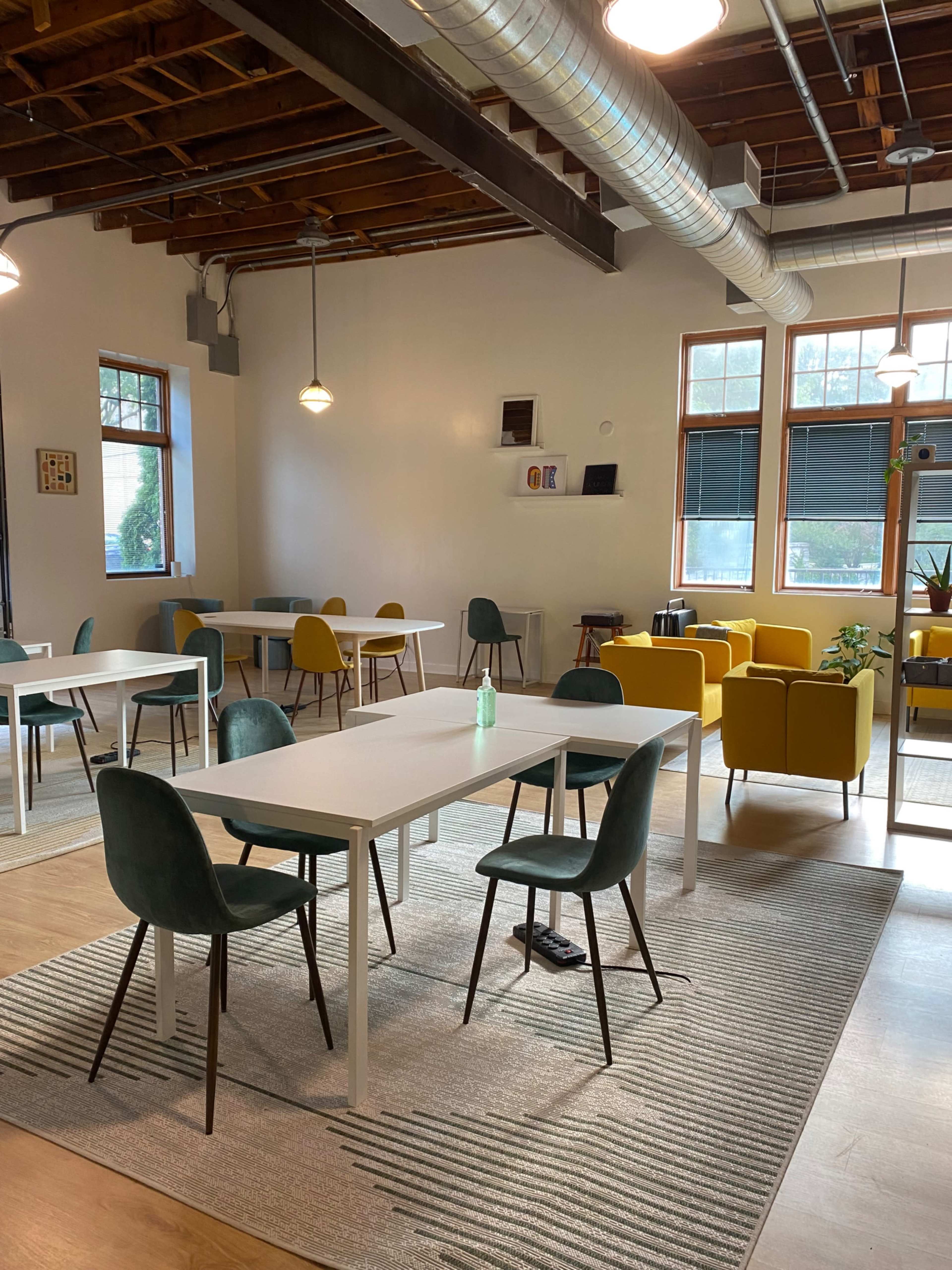 A brightly lit, modern workspace with several white tables surrounded by chairs in various colors, and a cozy seating area with yellow chairs.
