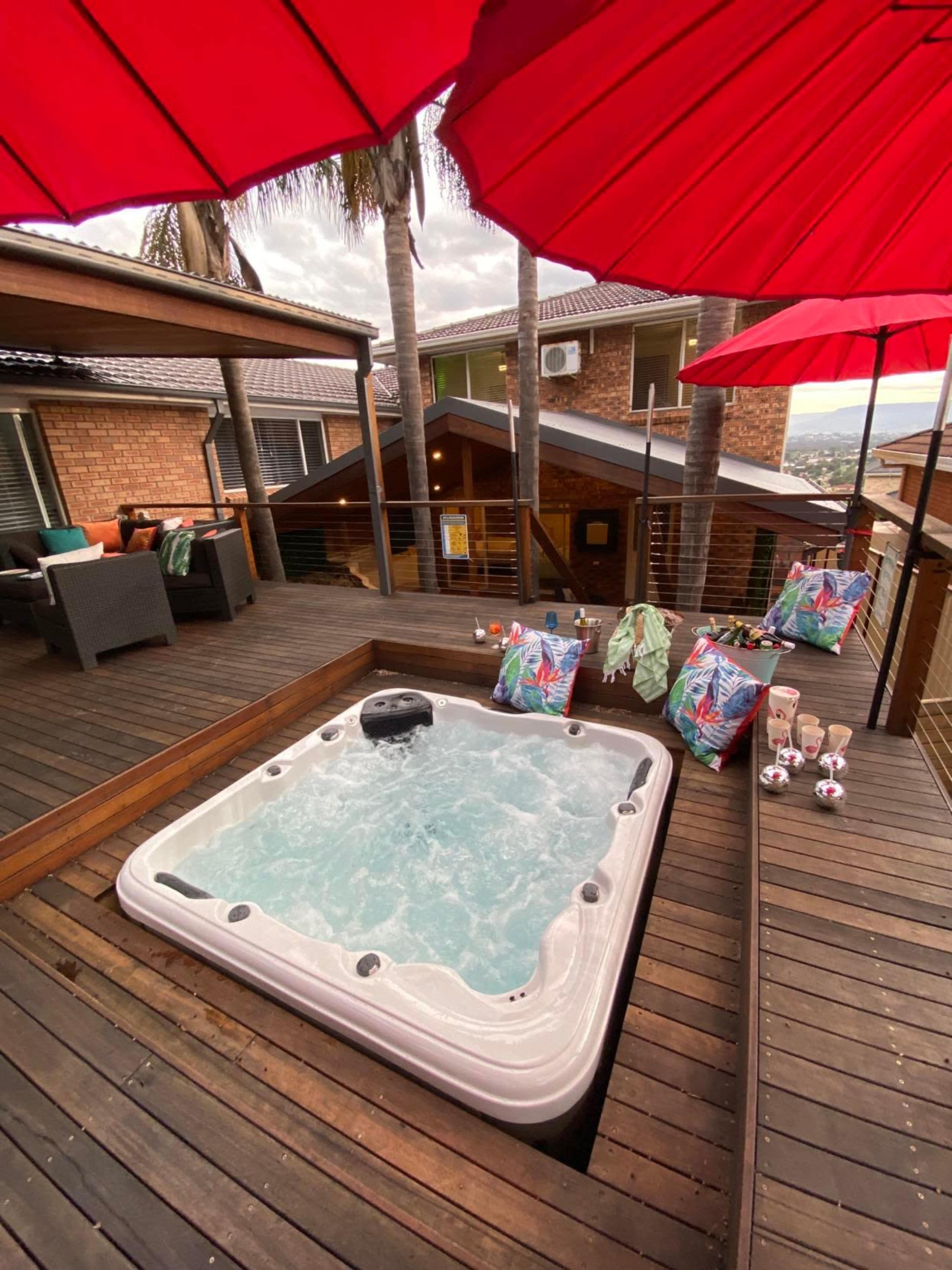 A hot tub is situated on a wooden deck surrounded by red umbrellas and lounge chairs, with various colorful towels and drinks arranged nearby.