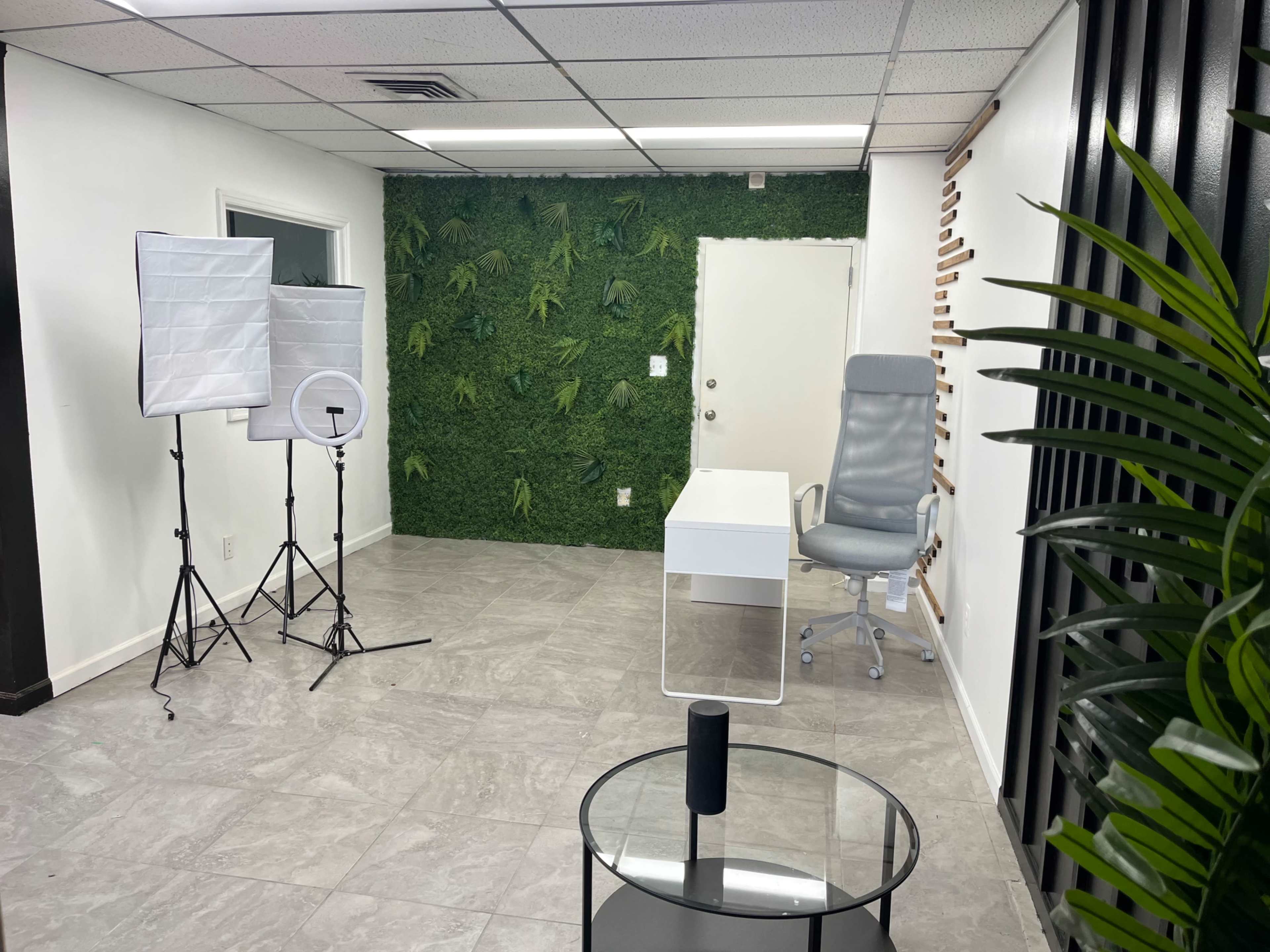 A modern office space with a white desk, a swivel chair, a green plant wall, and photography equipment set up in the corner.