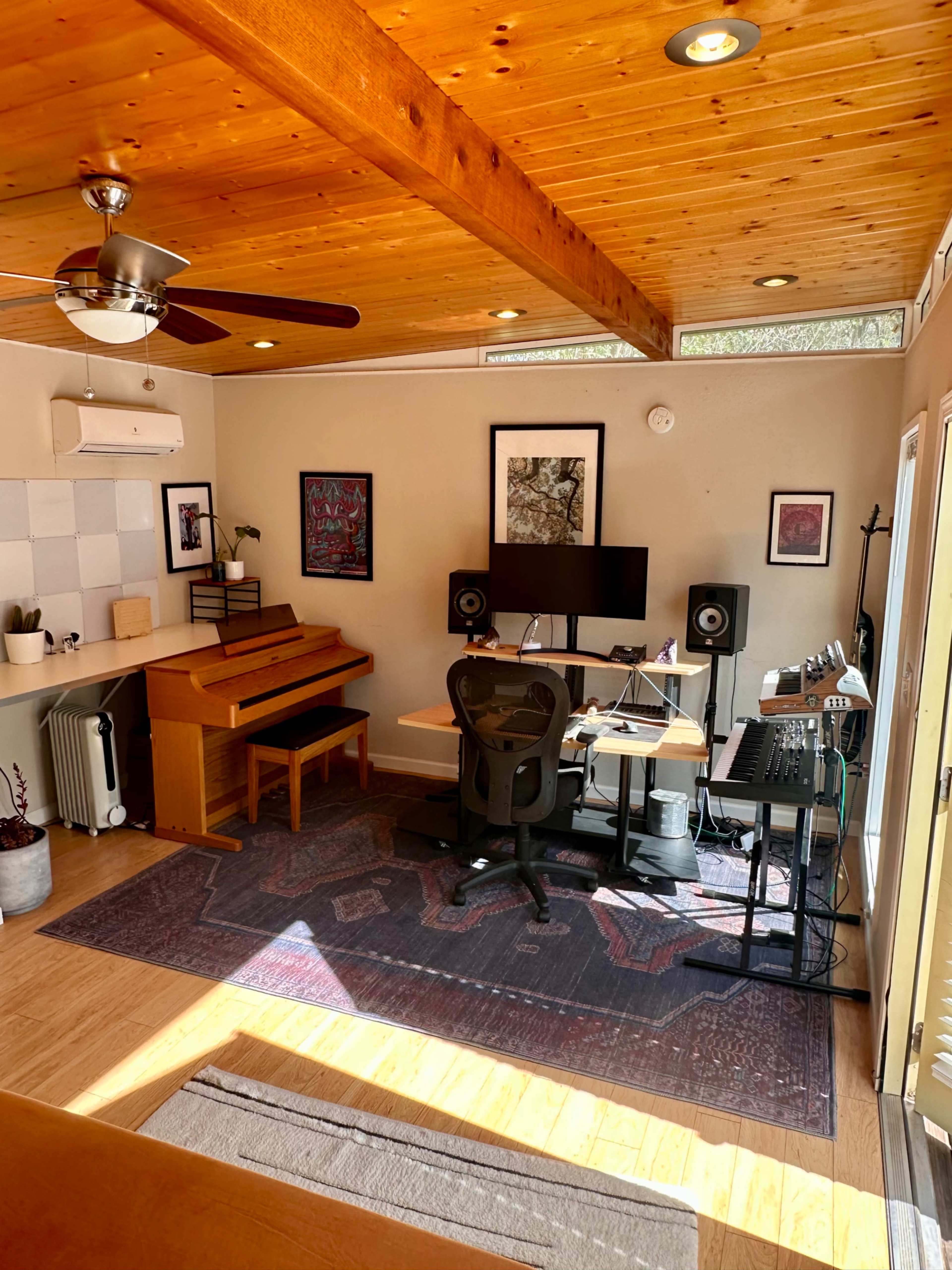 The image shows a bright room featuring a piano, a desk with a computer and audio equipment, and large windows allowing natural light to enter.
